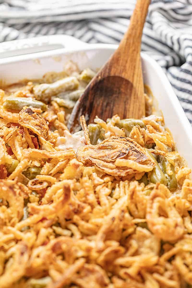 A wooden spoon dips into a pan of Easy Green Bean Casserole, topped with crispy fried onions.