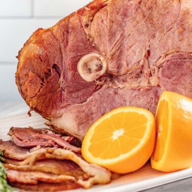 Angled view of full platter of a bone-in ham with slices laid out and garnished with sprigs of rosemary and orange halves.