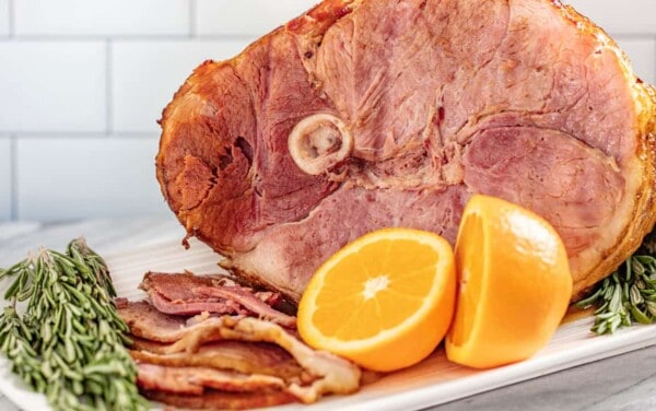 Angled view of full platter of a bone-in ham with slices laid out and garnished with sprigs of rosemary and orange halves.