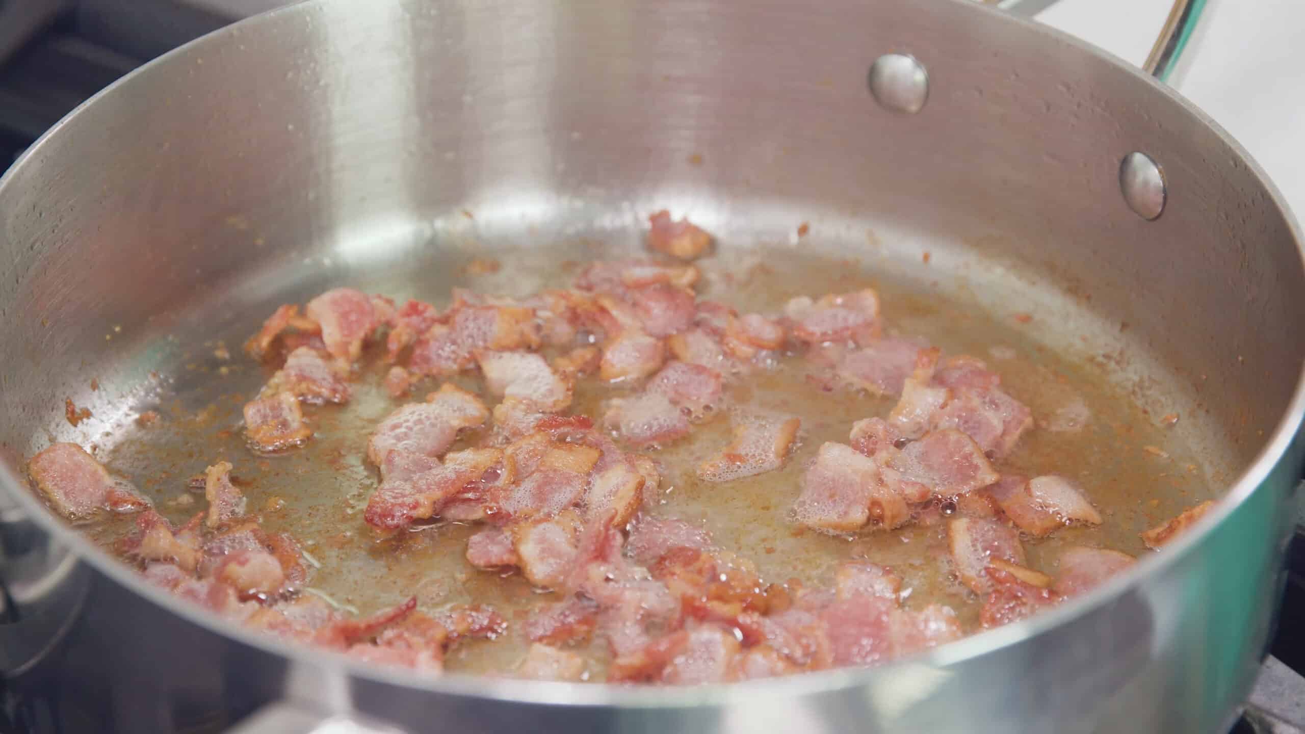 Close-up view of interior of stainless steel sauce pan with chopped pieces of bacon sizzling inside.