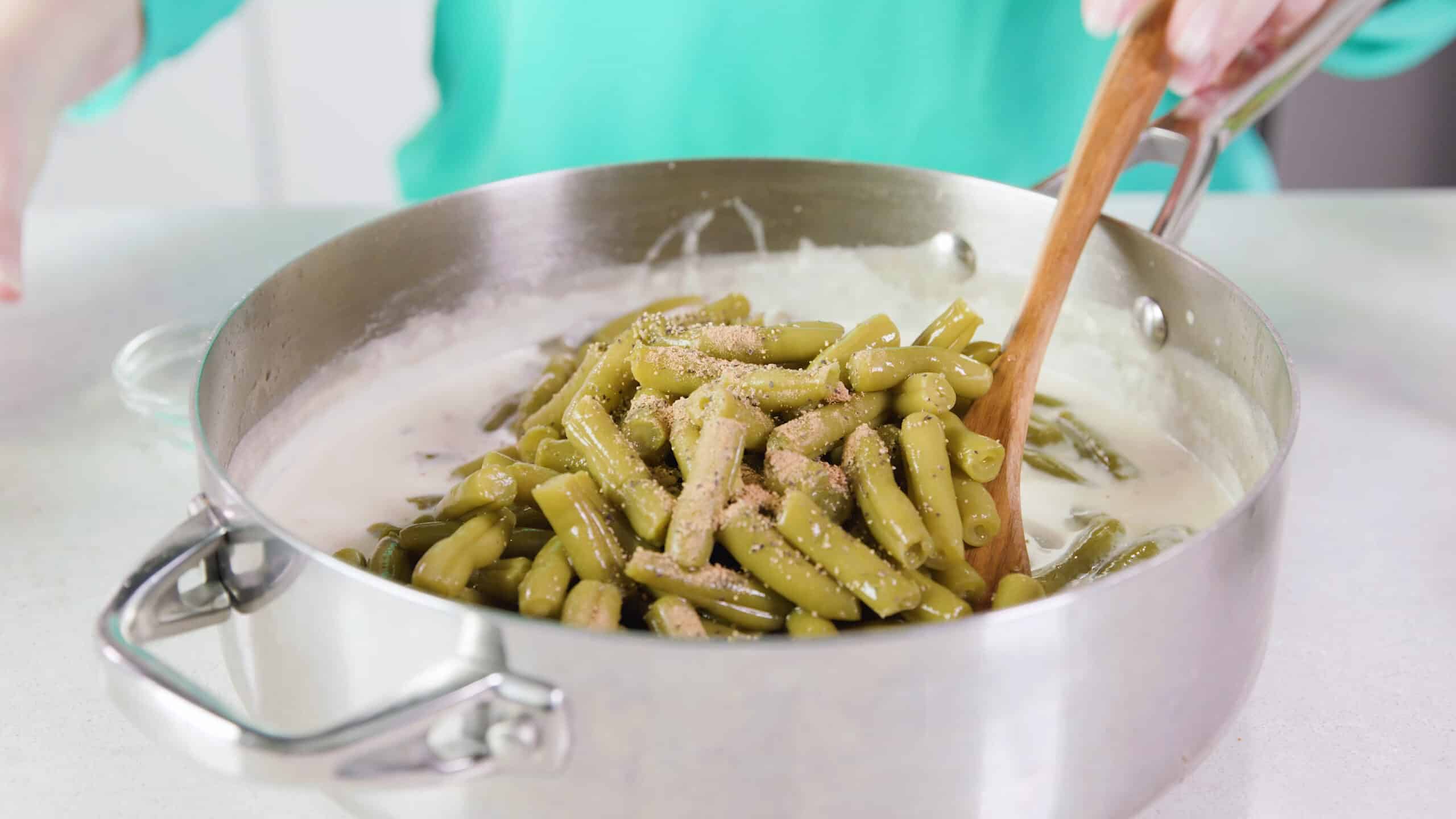 Angled view of high-rim stainless steel saucepan filled with creamy sauce and added green beans cover in various spices and a wooden spoon ready to mix.
