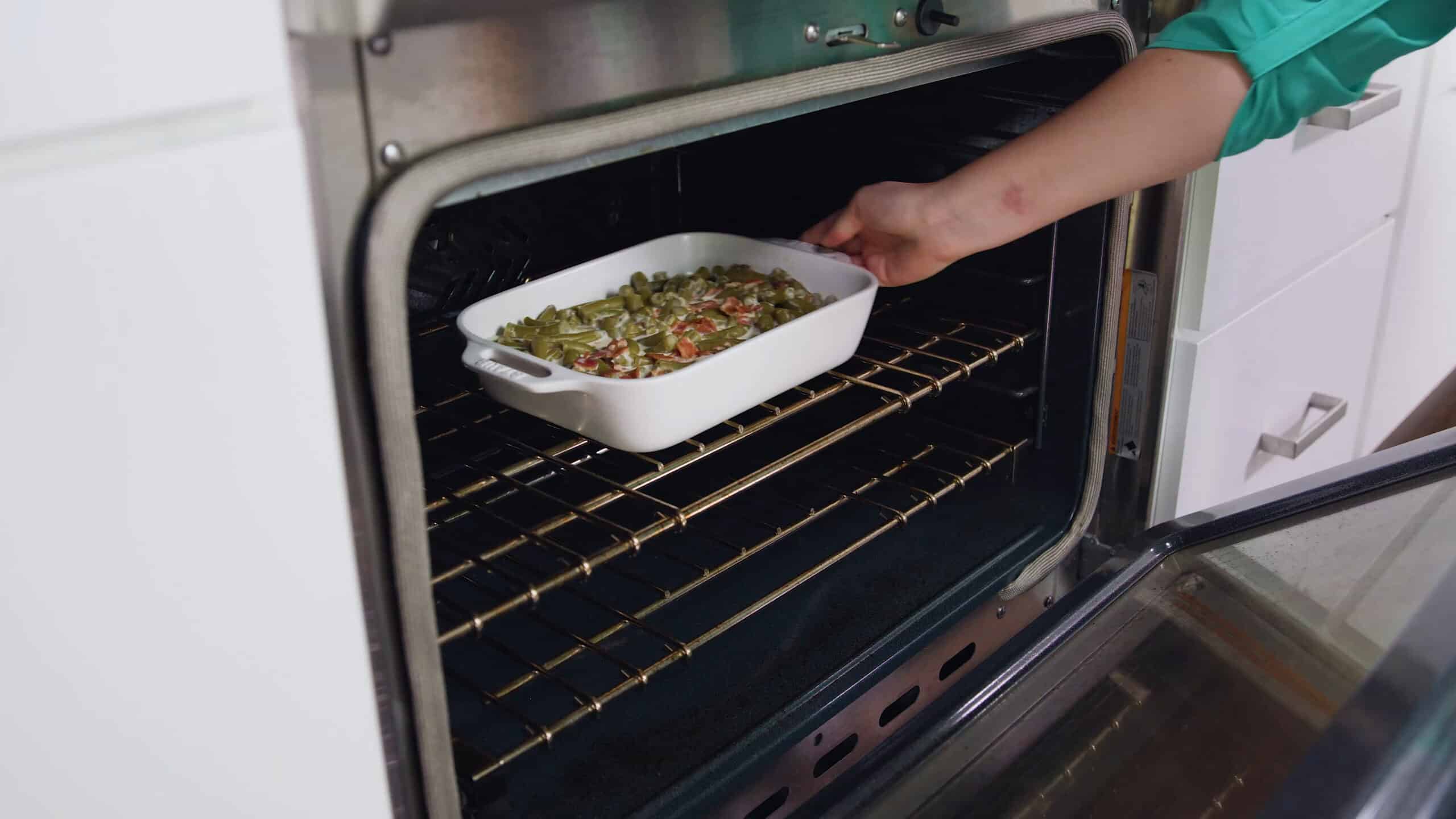 Angled view of open oven with white casserole dish filled with green bean casserole filling, placed on rack in the middle of the oven.