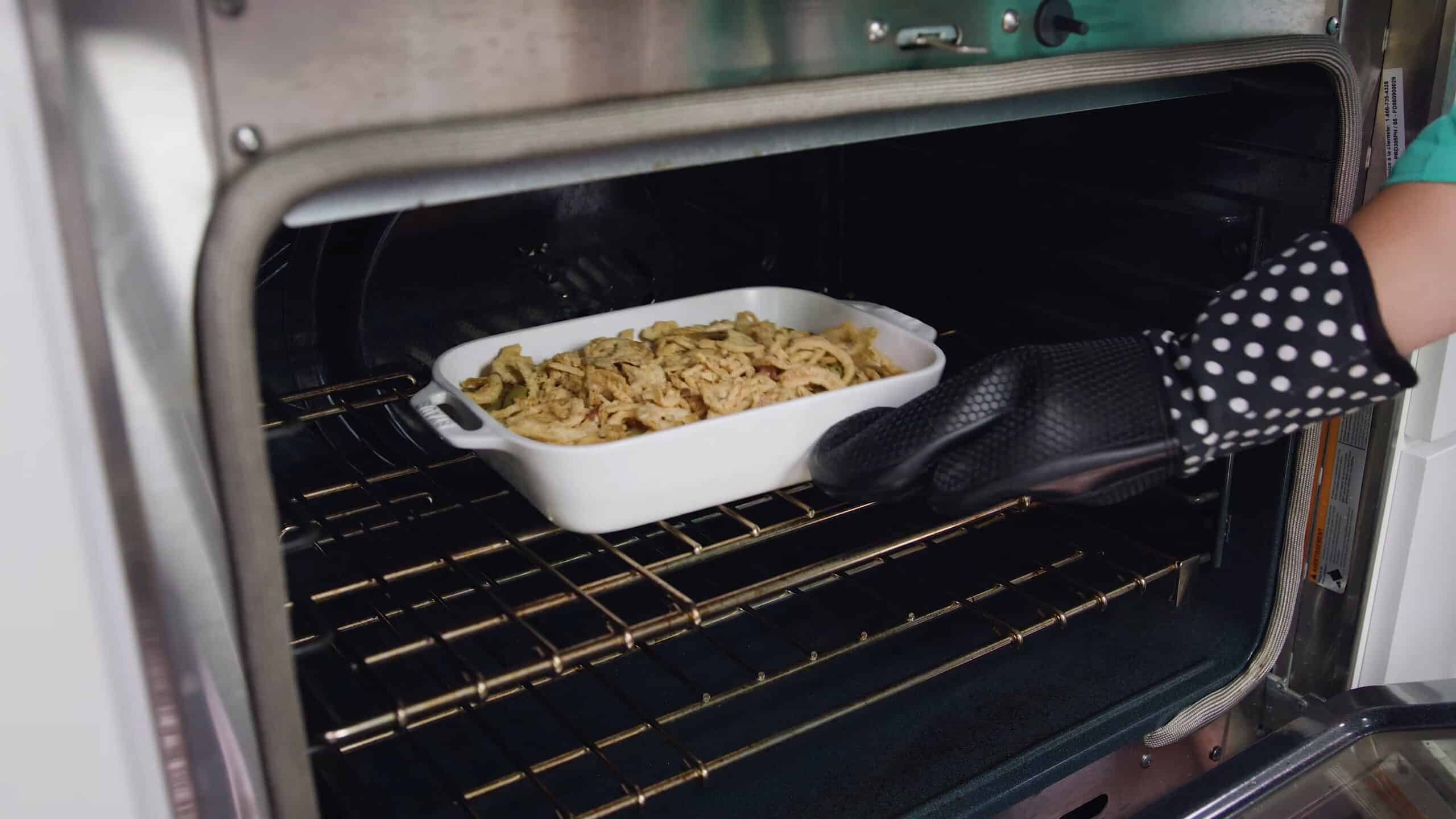 Angled view of open oven with white casserole dish filled with green bean casserole filling and topped with French fried onions, placed on rack in the middle of the oven.