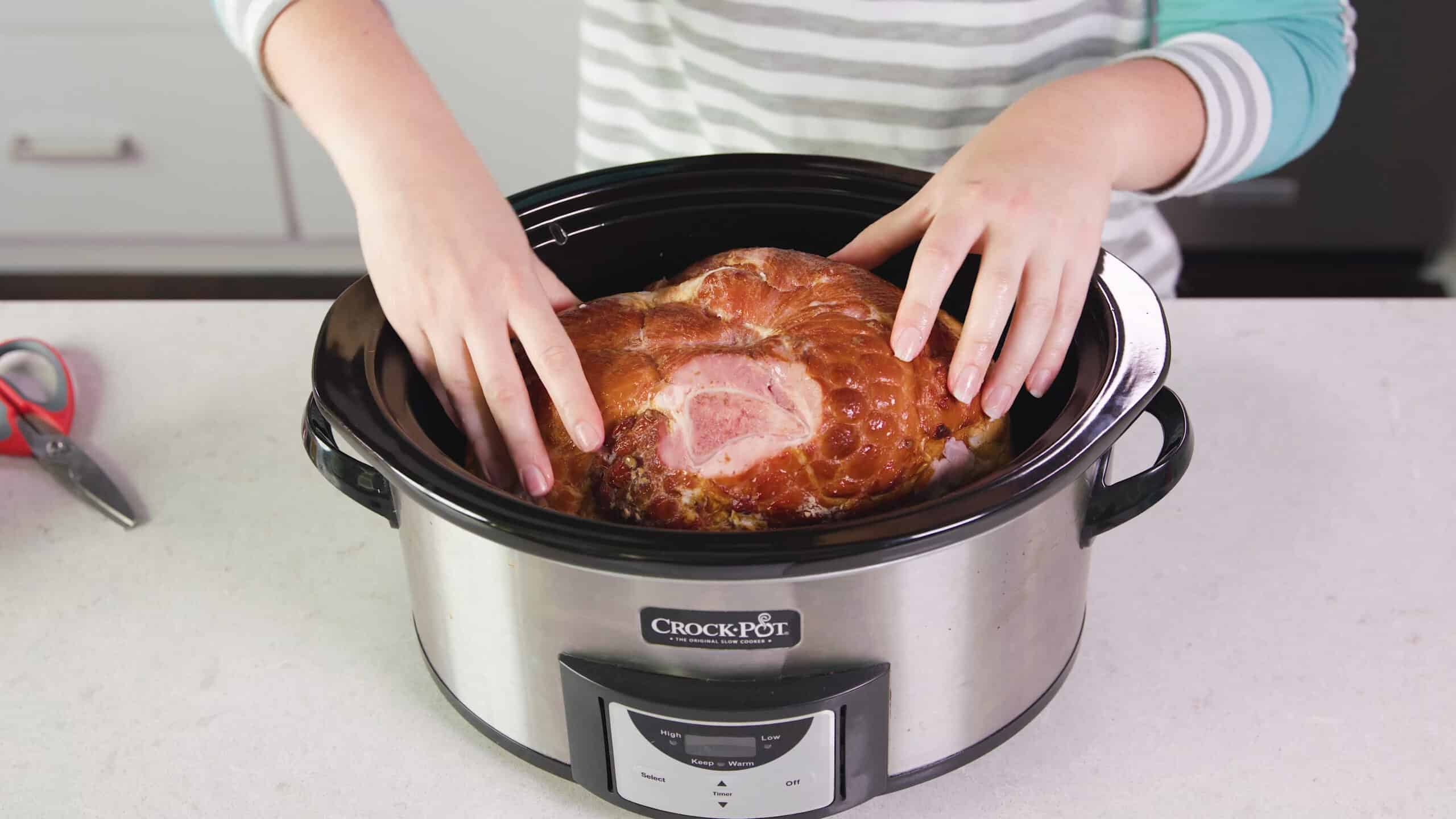 Angled view of slow cooker with ham inside on marble countertop.