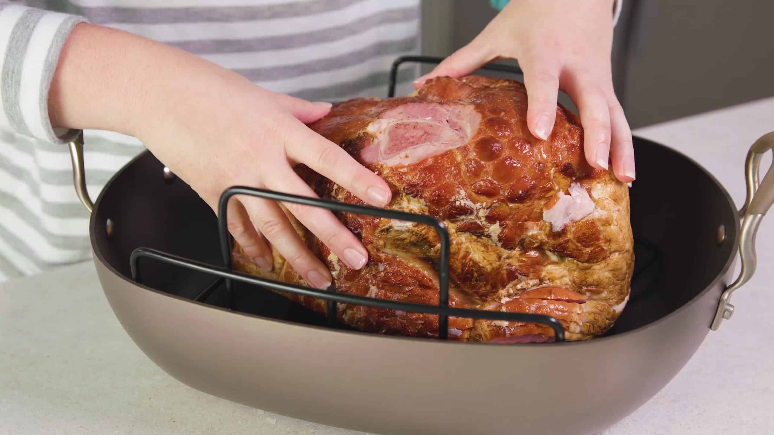 Angled view of stainless steel roasting pan with metal rack to lift the ham that is inside above the bottom of the pan.
