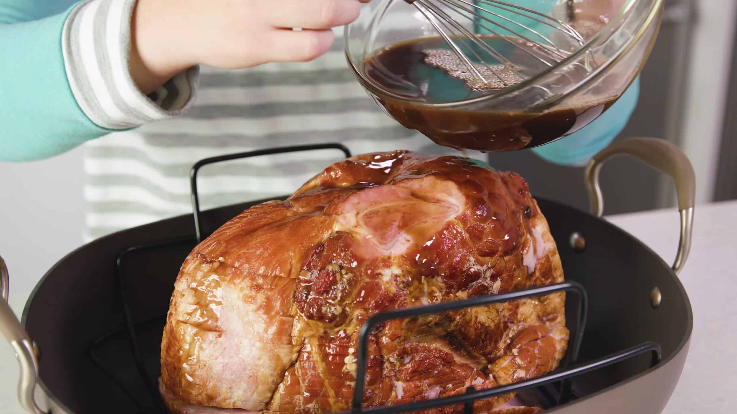 Angled view of stainless steel roasting pan with rack and ham inside and a large glass mixing bowl with orange sauce glaze being pour onto the ham using a wire whisk to guide the liquid out.