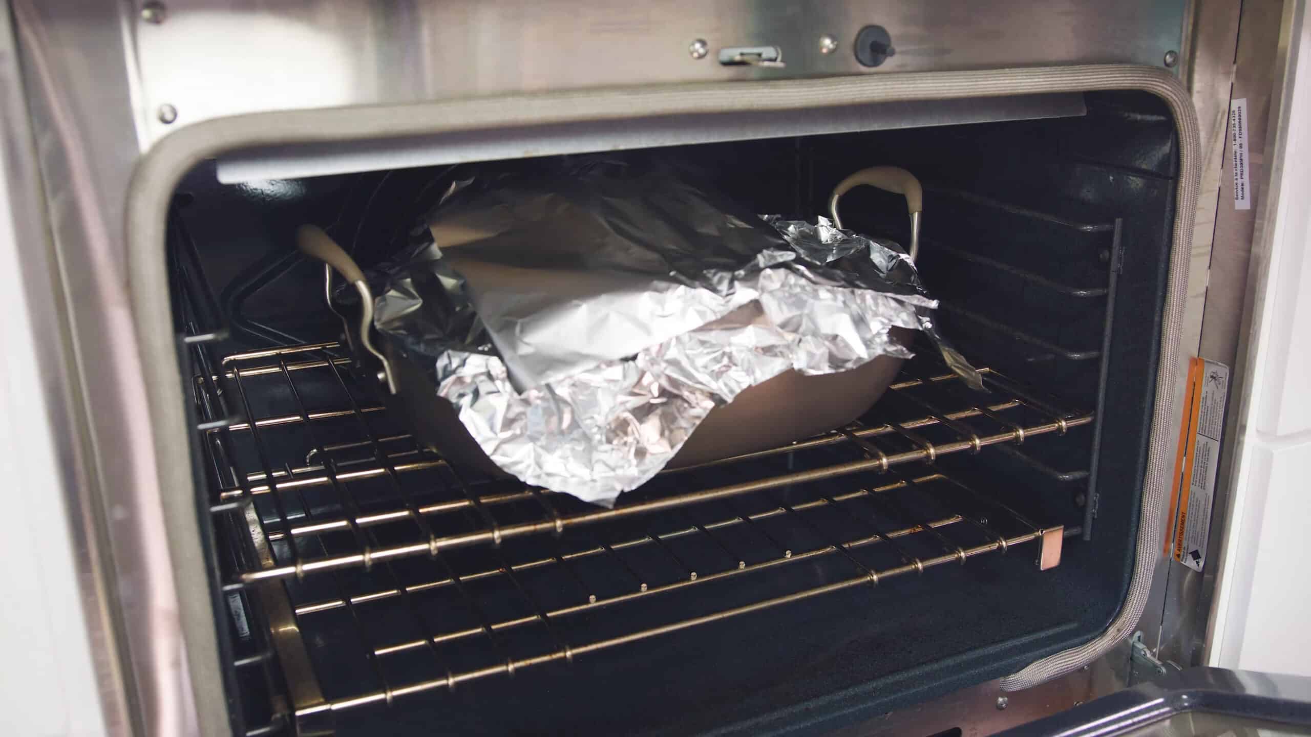 Angled view of open oven with stainless steel roasting pan covered in aluminum foil on rack in the middle of the oven.