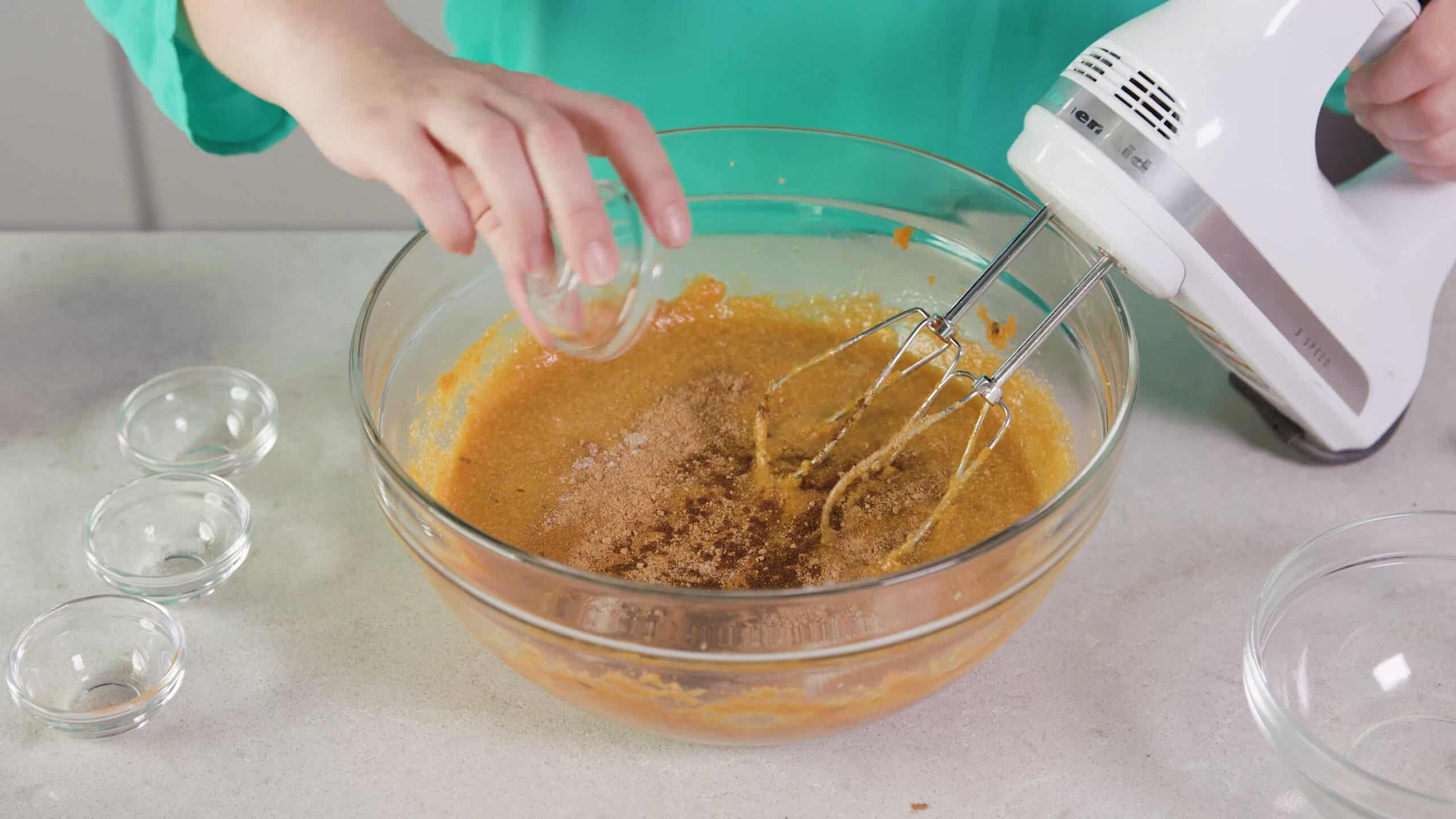 Angled view of large clear glass mixing bowl filled with mashed and mixed sweet potatoes and other spice ingredients with a hand mixer at the ready.