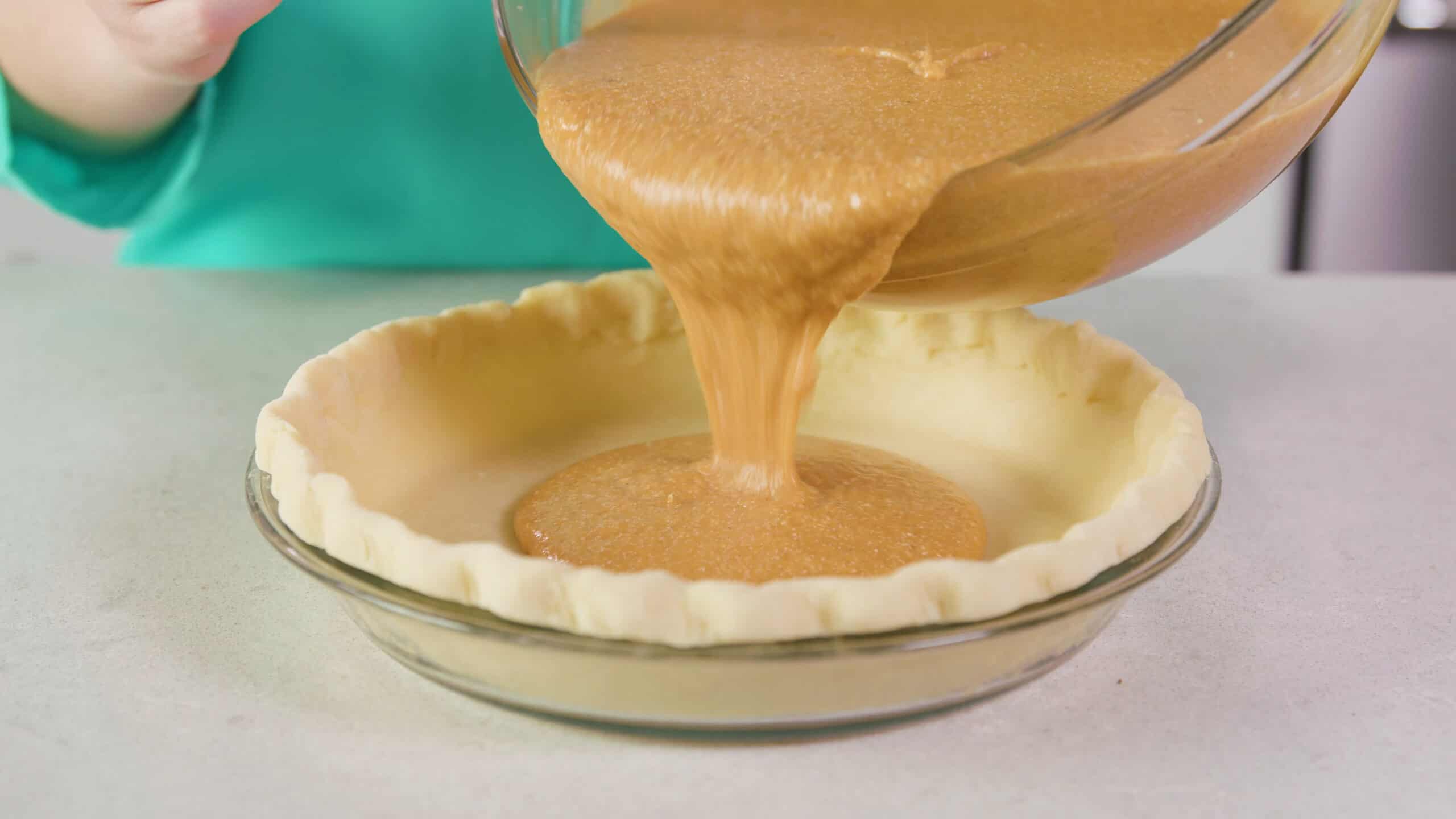 Angled view of prepared uncooked pie crust in a clear glass pie plate with sweet potato pie filling being poured in from large clear glass mixing bowl.