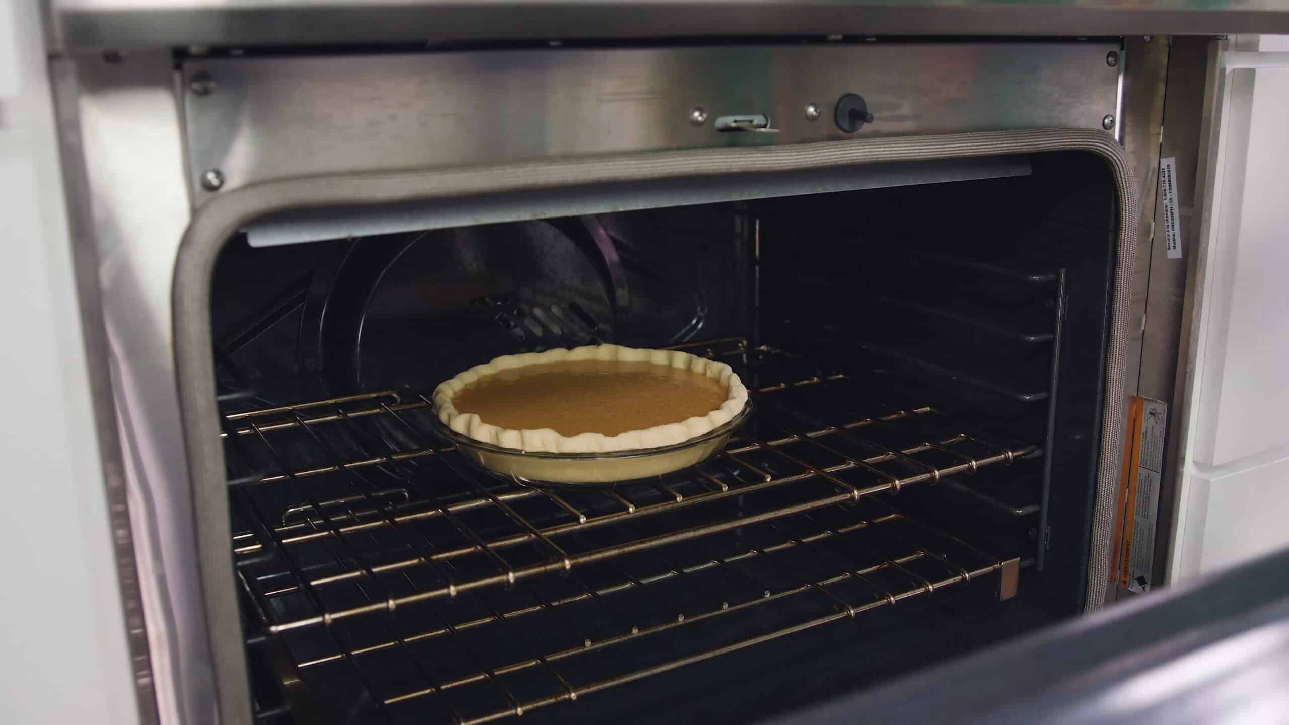 Angled view of open oven with clear glass pie plate filled with uncooked pie crust filled with sweet potato pie filling on a rack in the middle of the oven.
