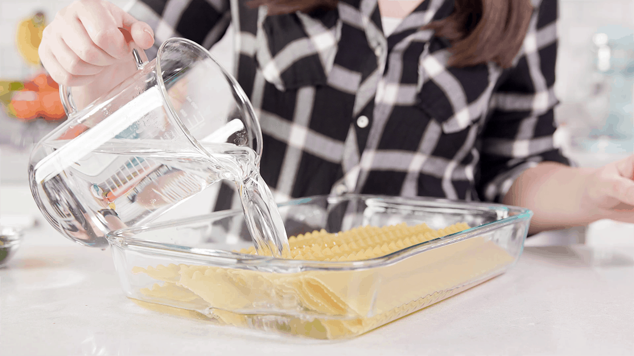 Water being poured over dry lasagna sheets in a clear glass 9x13 pan