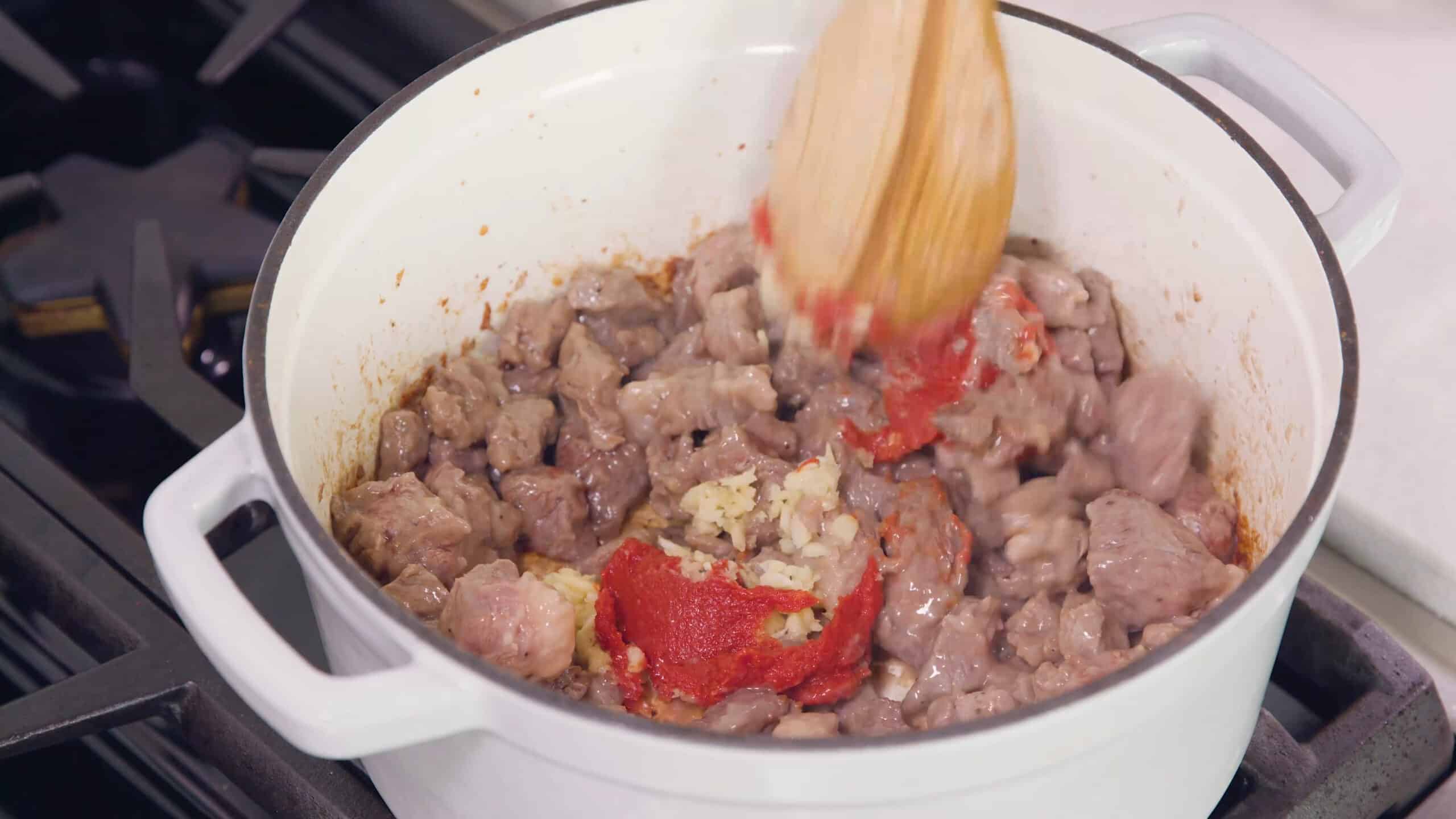 Angled view of white enamel cast iron pot filled with cooking beef chunks and added tomato paste and minced garlic with a wooden spoon ready to mix.