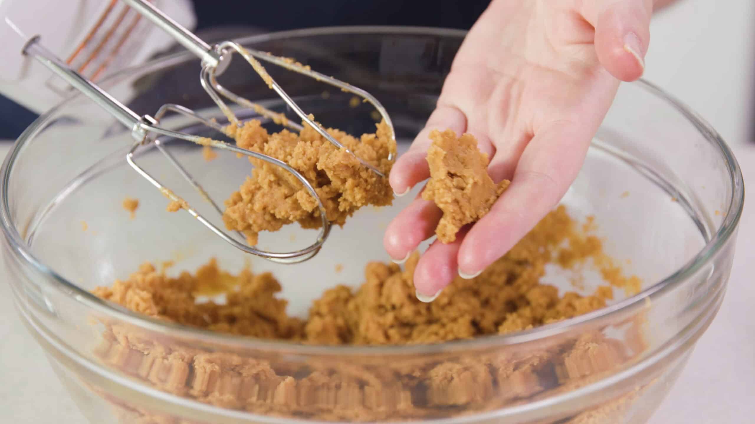 Close-up view of a hand with bit of pinched peanut butter cookie dough to show how the dough comes together to form cookie dough balls with hand mixer beaters covered with cookie dough and a clear glass mixing bowl filled with cookie dough in the background.