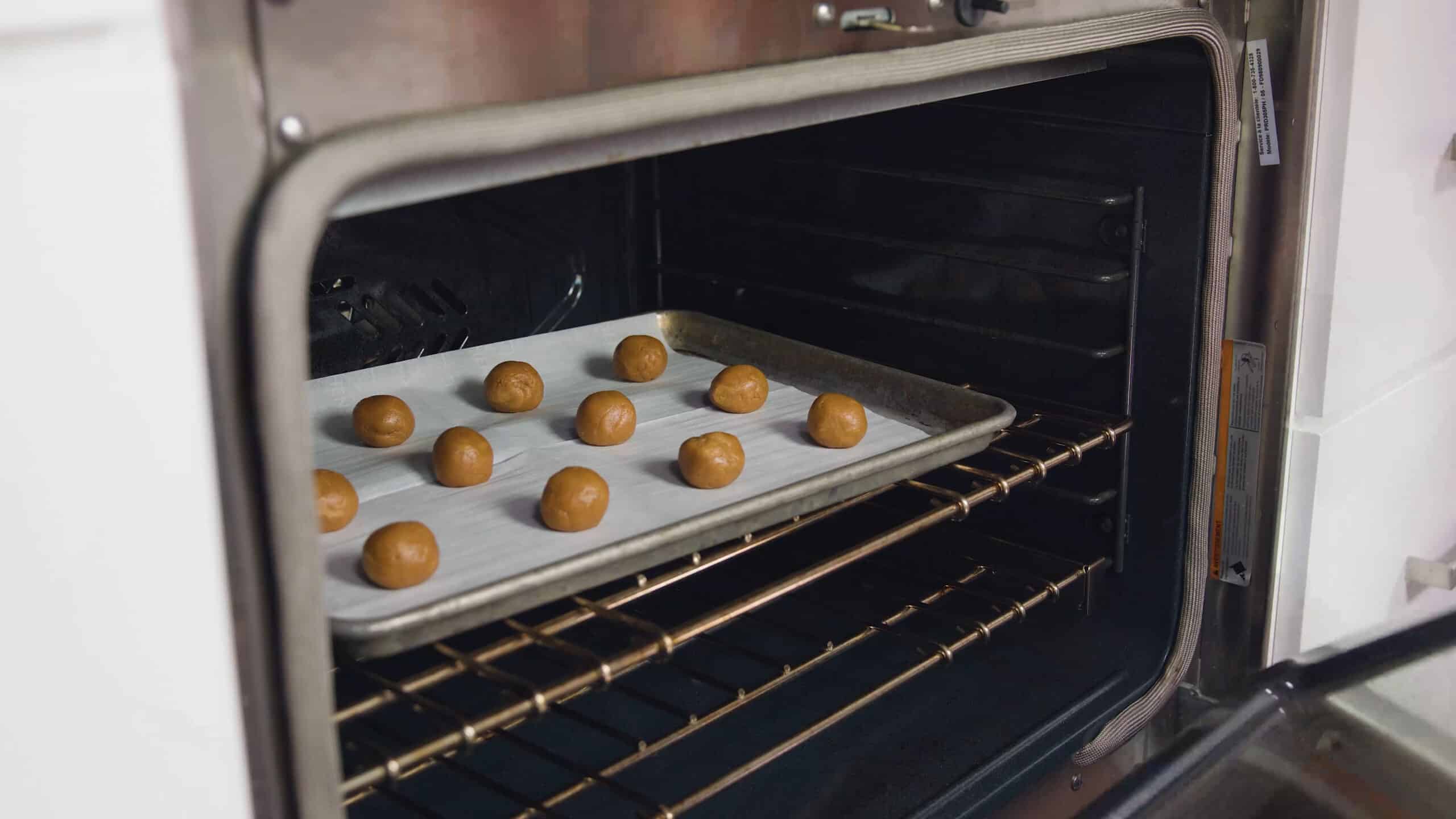 Angled view of open oven with silver baking sheet lined with parchment paper and one dozen peanut butter cookie dough balls on a metal rack in the middle of the oven.