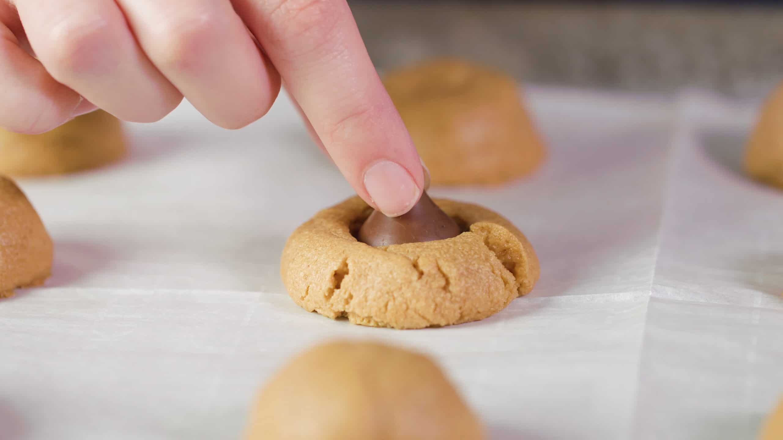 Close up view of one peanut butter cookie in the middle of a parchment lined cookie sheet and a hand pushing a chocolate kiss candy into the cookie on top.