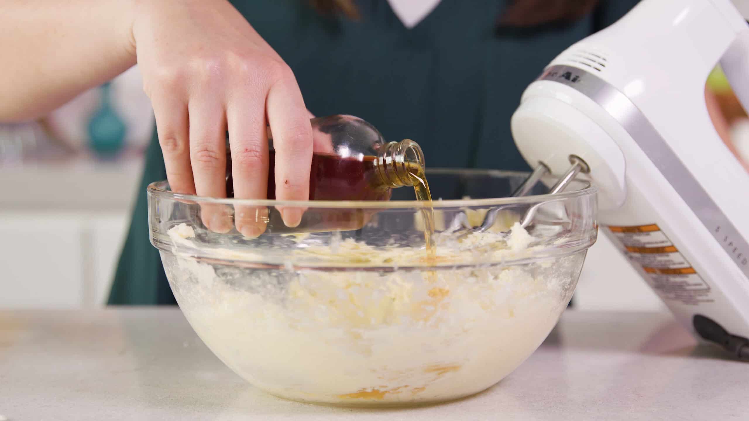 Side view of a large clear glass mixing bowl filled with cookie batter and a hand pouring vanilla extract from a clear glass bottle and a hand mixer leaning in the bowl in the background.