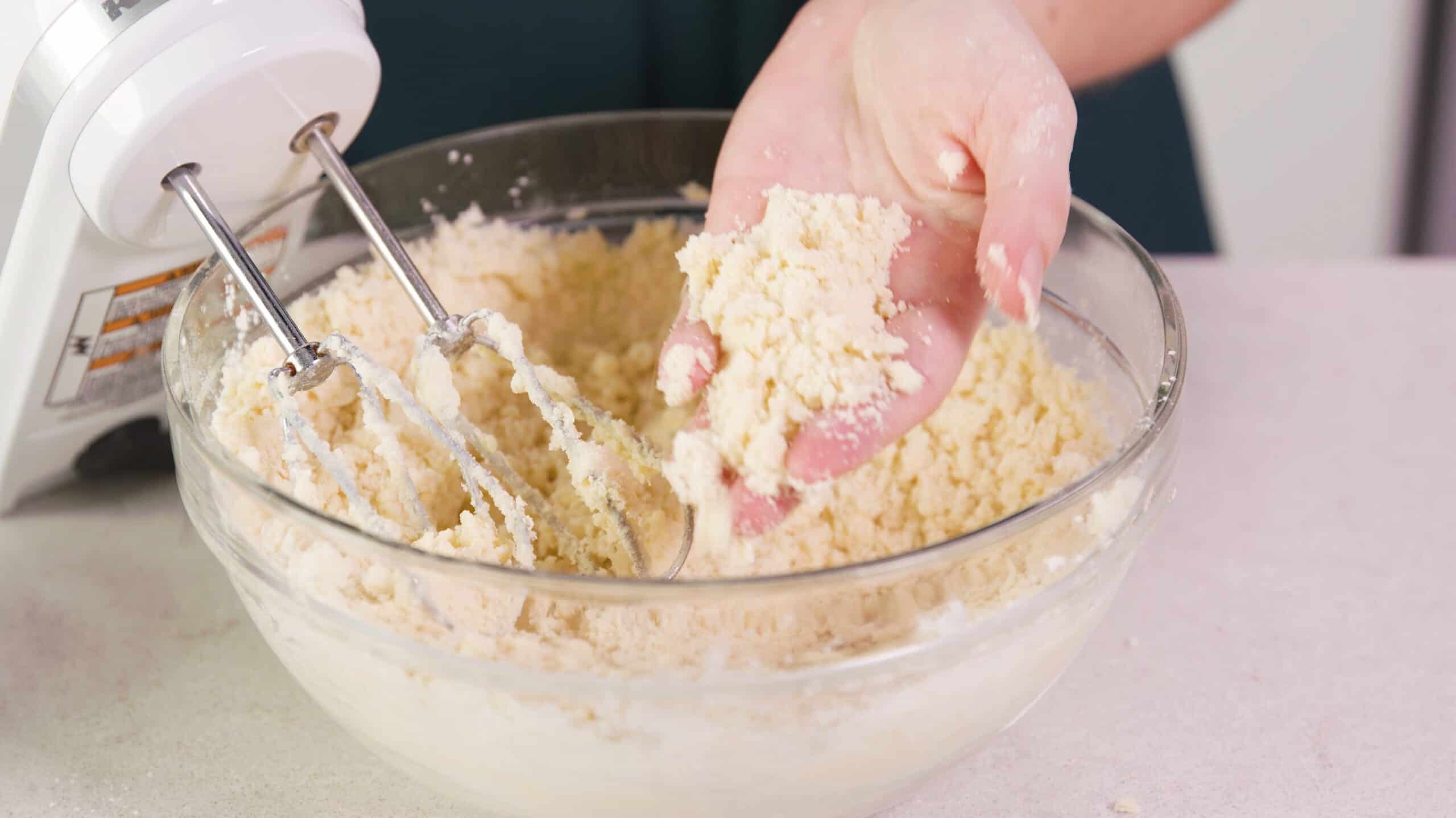 Close up view of large clear glass mixing bowl filled with cookie batter and a hand filled with batter to show a sand-like texture with a hand mixer leaning into the bowl with metal beaters.