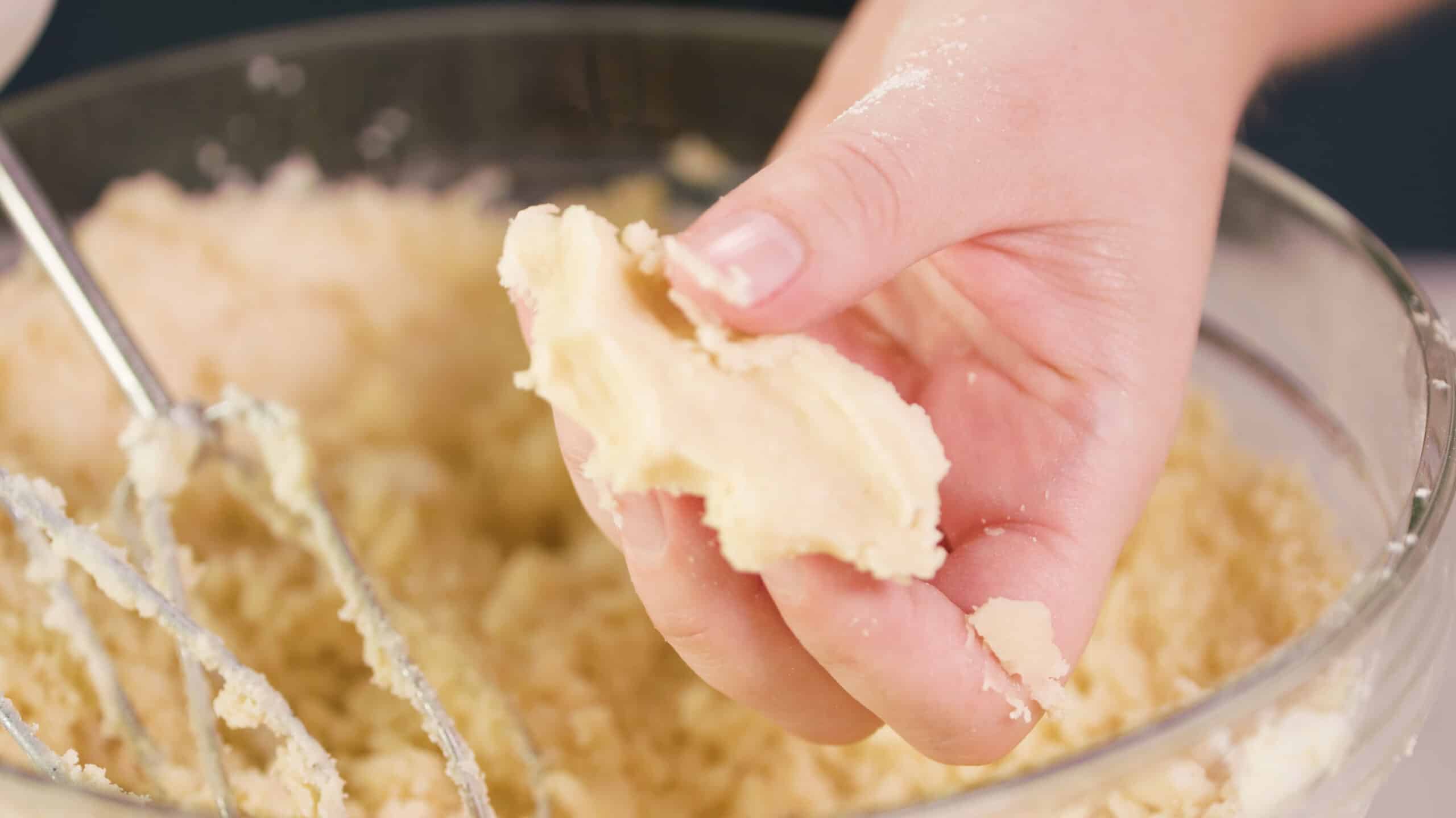 Close up view of a hand holding squeezed cookie batter to show the desired texture when the batter is pressed with a large clear glass mixing bowl filled with cookie batter in the background with metal beaters leaning into the bowl from a hand mixer.