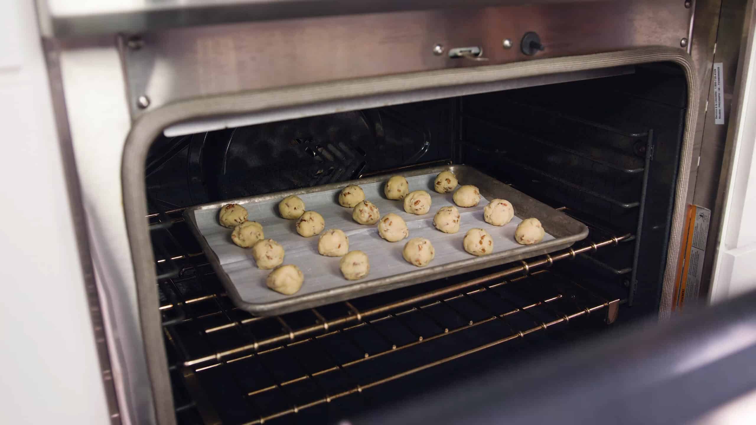 Angled view of an open oven with a metal baking sheet lined with parchment paper and twenty rolled pecan sandies placed on top all on a metal racking the middle of the oven.