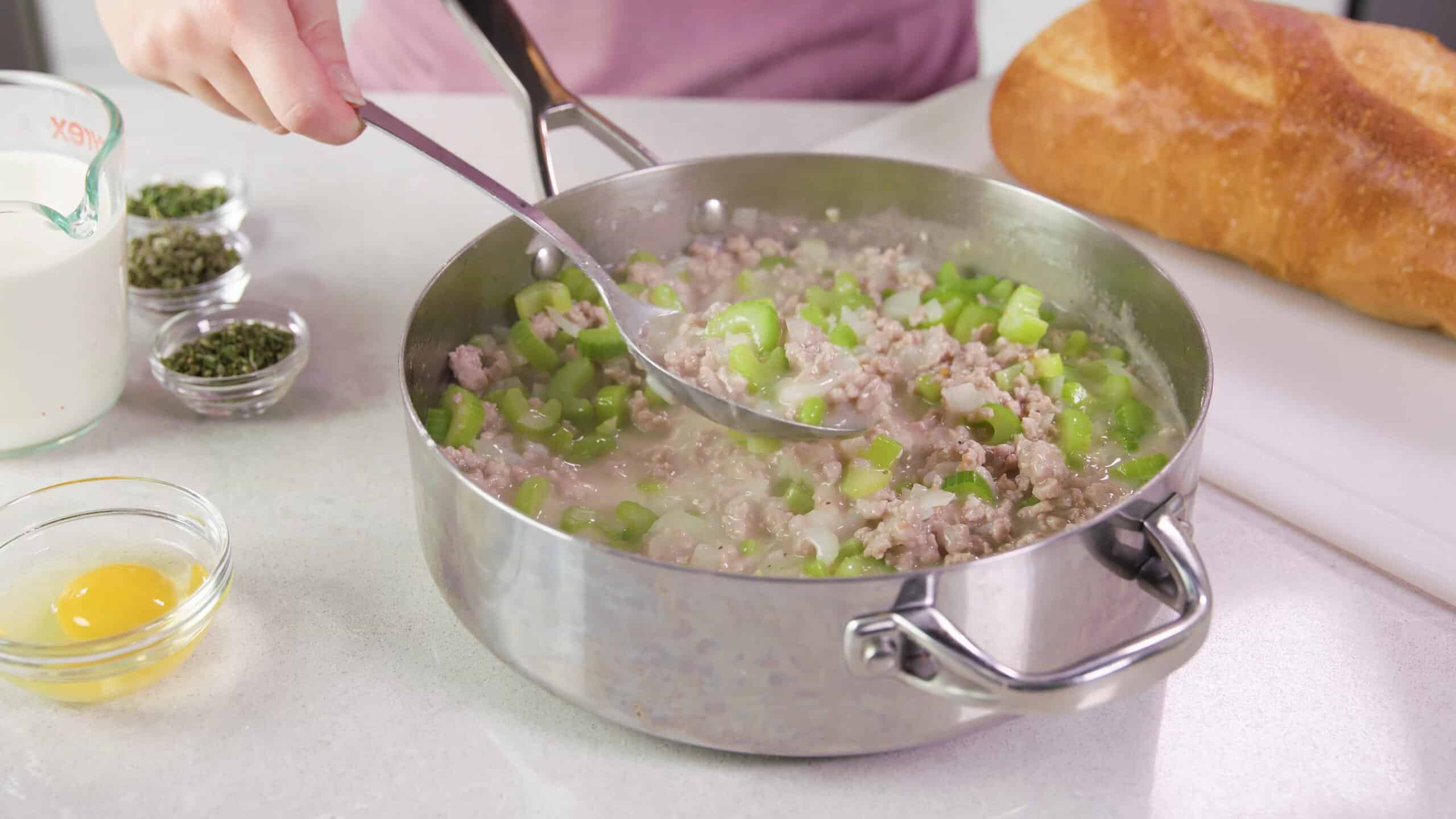 Angled view of high-rim saucepan filled with sausage, celery, onion and seasoning mixture with a metal spoon lifting a portion up to show texture.
