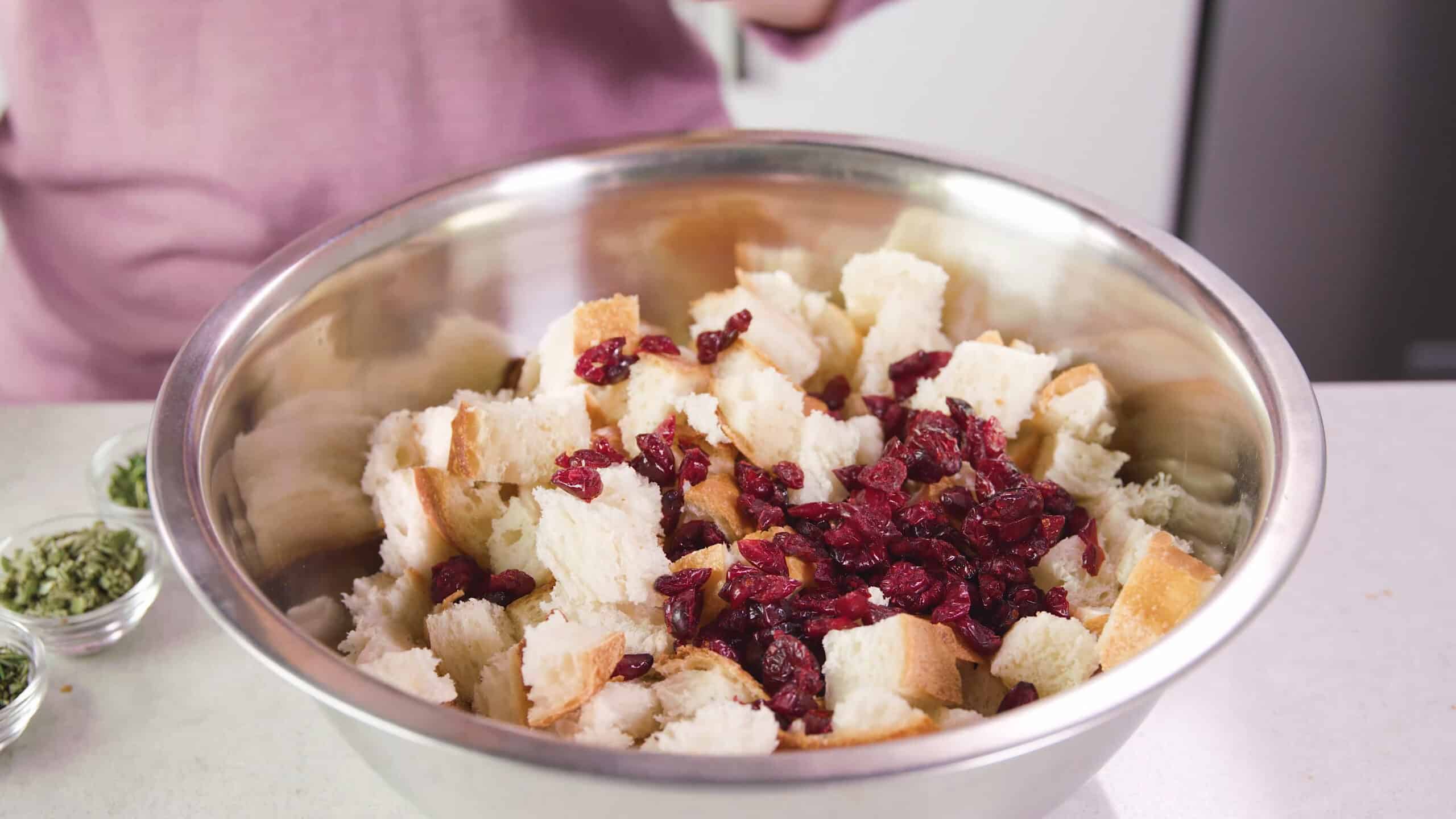 Angled view of stainless steel mixing bowl filled with diced bite-sized portions of French bread covered with dried cranberries.
