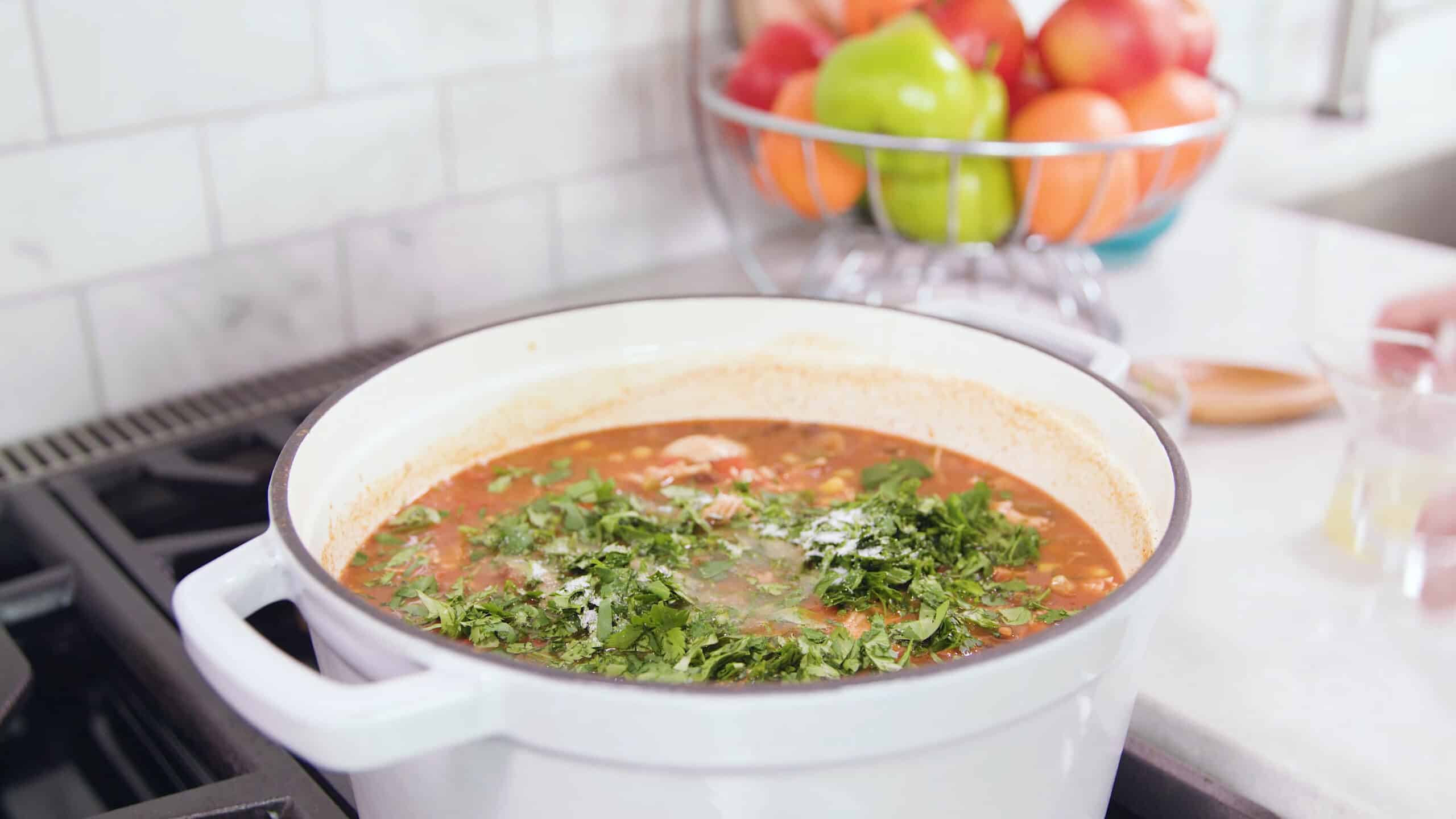 Angled view of white enamel coated cast iron pot filled with soup base and topped with chopped cilantro and salt to taste.