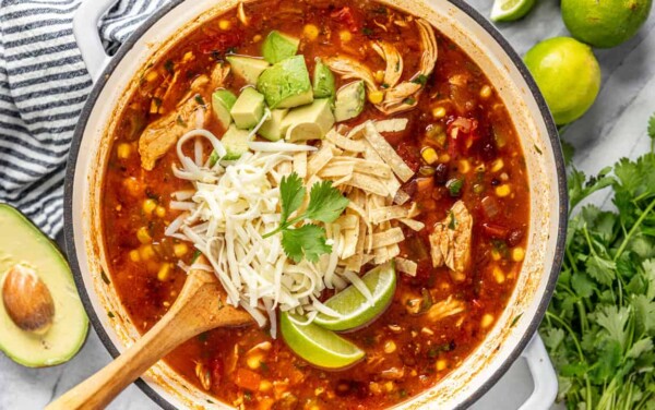 Bird's eye view of chicken tortilla soup topped with cheese, avocado, tortilla strips, lime, and cilantro, served out of a large dutch oven pot with a wooden spoon.