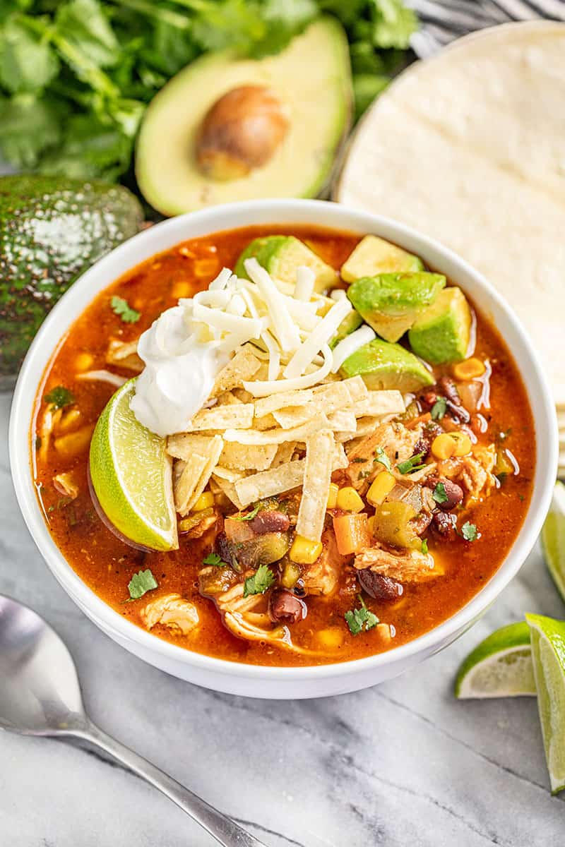 Bowl of chicken tortilla soup topped with cheese, avocado, tortilla strips, lime, and cilantro