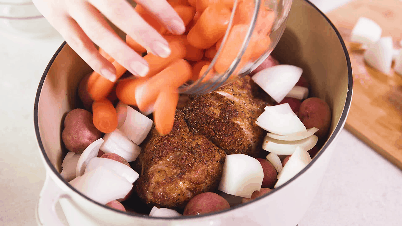 Carrots being scattered over a pork roast surrounded by onions and potatoes in a dutch oven pot