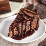 A slice of Chocolate Cheesecake with Oreo Crust, served on a plate and drizzled with chocolate syrup.