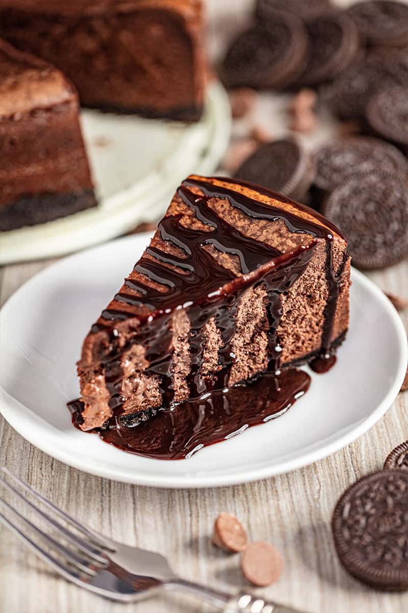 A slice of Chocolate Cheesecake with Oreo Crust, served on a plate and drizzled with chocolate syrup.