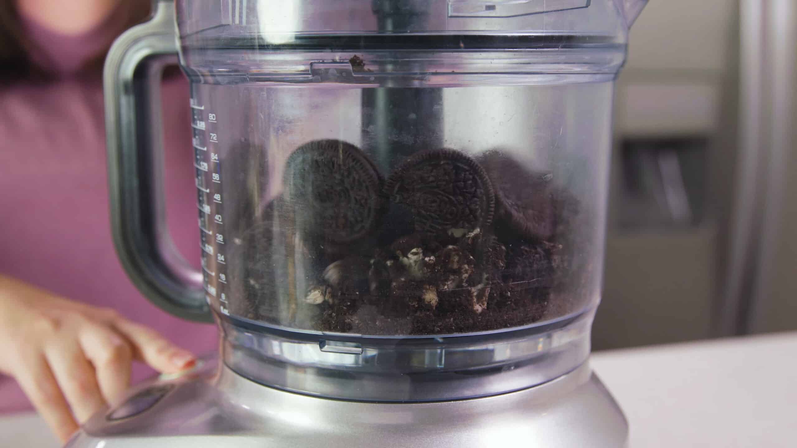 Side view of a food processor filled with Oreo cookies and pulsing to finely chop the cookies.