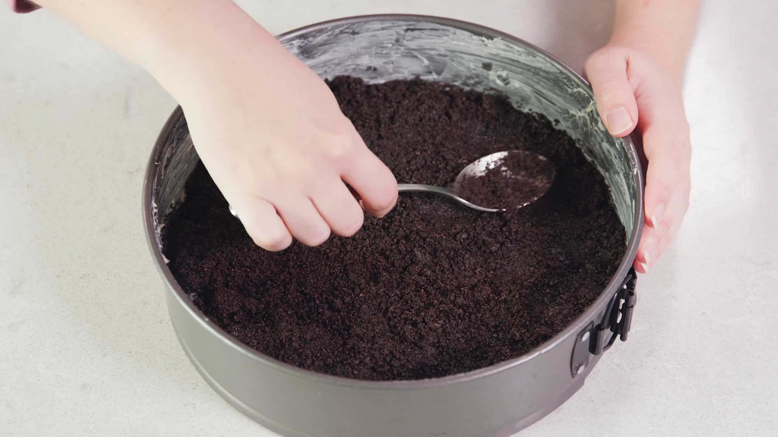 Overhead view of metal spring form pan greased with a stick of butter and the bottom coated in crushed Oreo cookies and pressed down with a metal spoon.