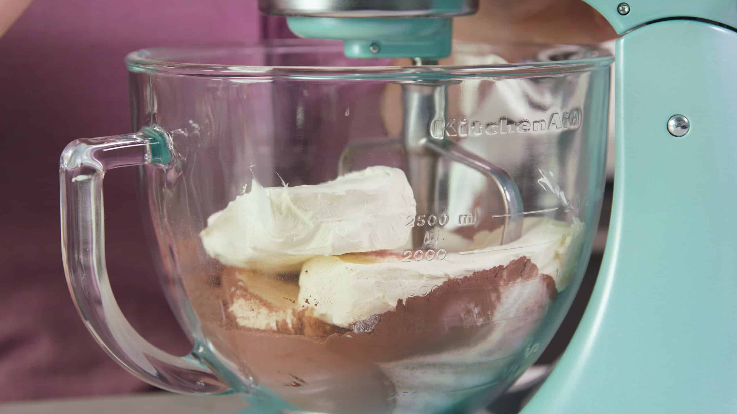 Side view of clear glass measuring bowl connected to a countertop mixer with a metal mixing paddle filled with cocoa powder, cream cheese and other batter ingredients.
