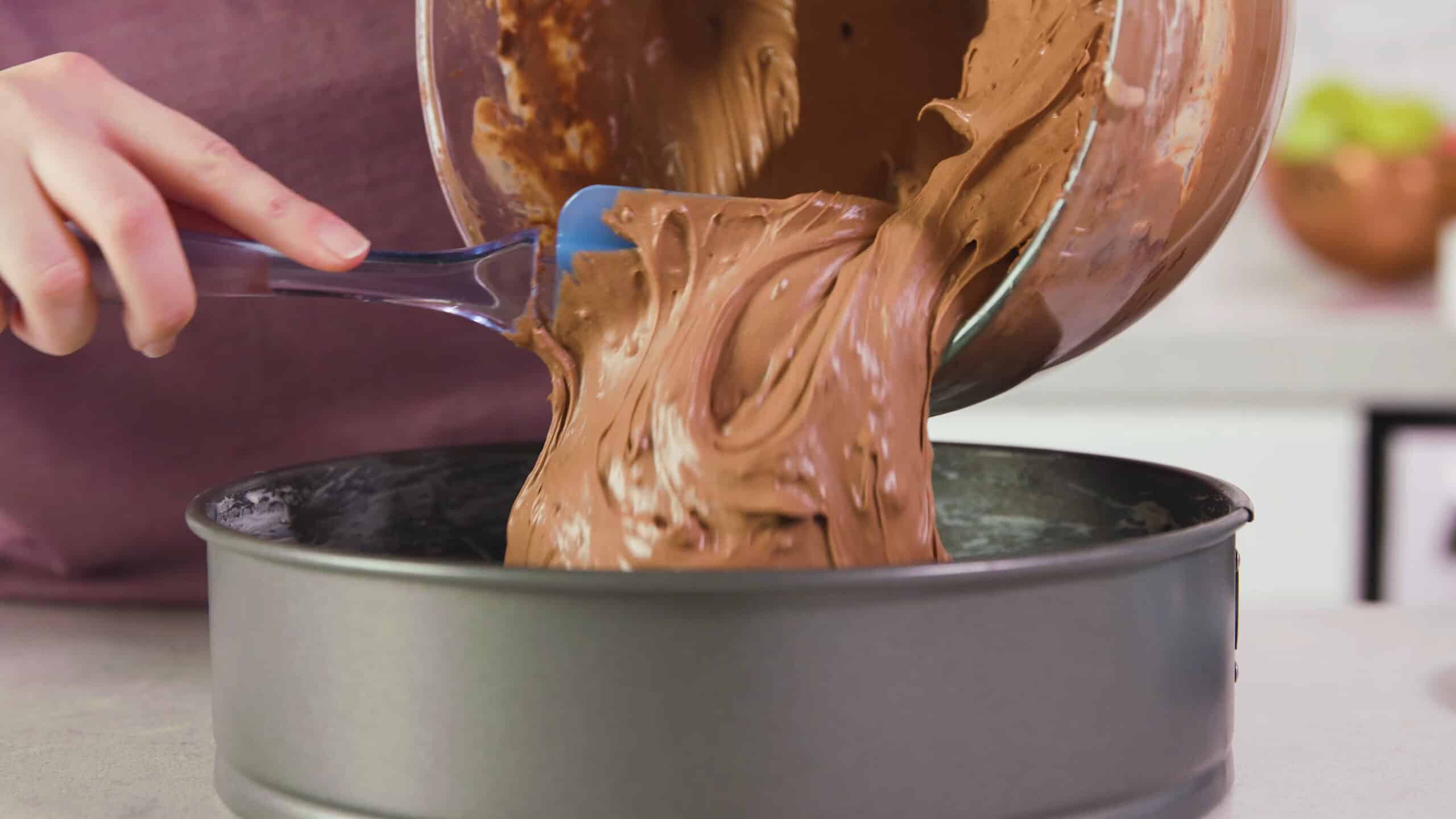 Side view of metal spring form pan being filled with chocolate cheesecake batter from a clear glass mixing bowl scooped out with a plastic spatula.
