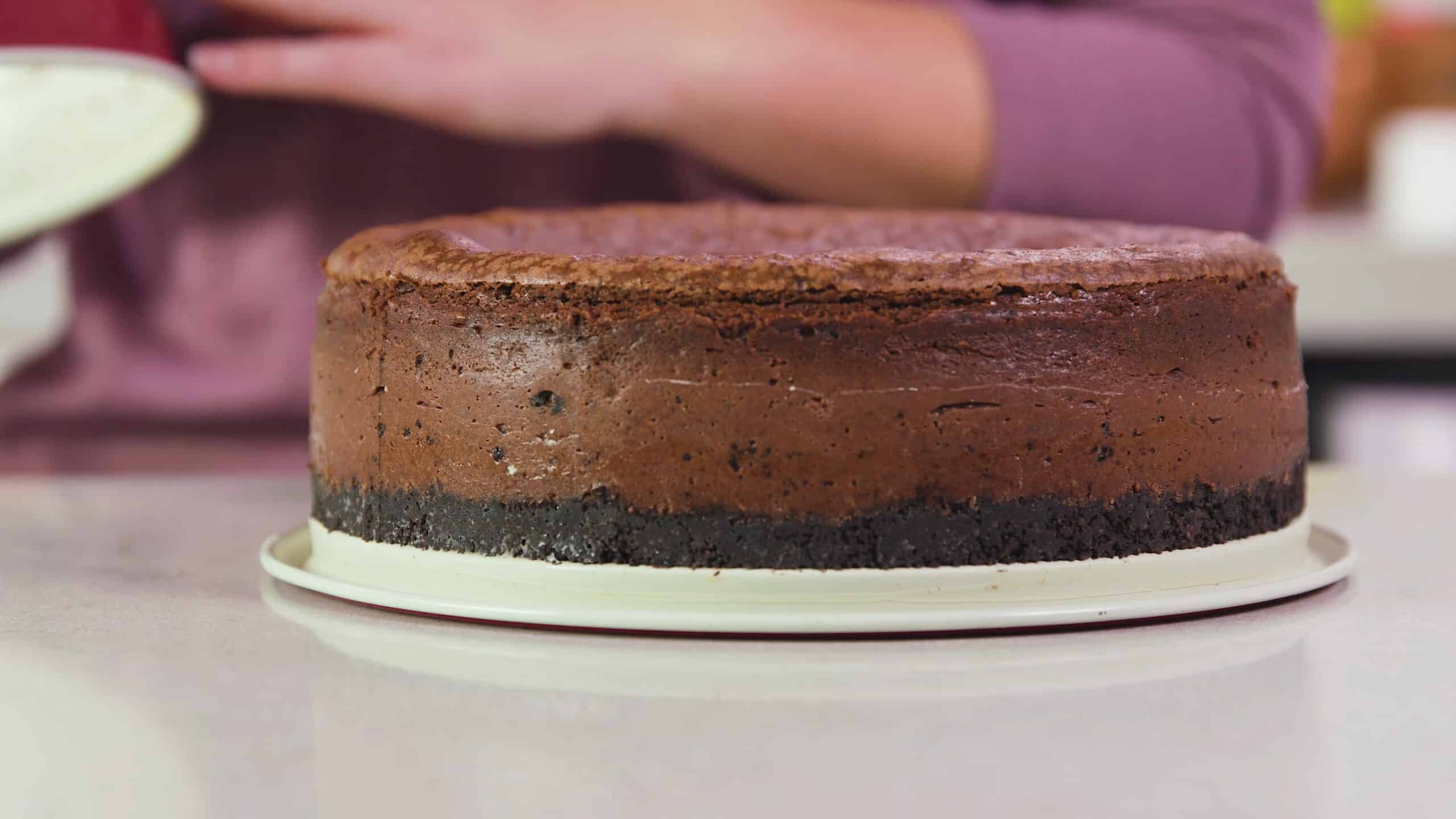 Side view of completely baked chocolate cheesecake with Oreo crust resting on the bottom part of the metal spring form pan after the side has been removed.