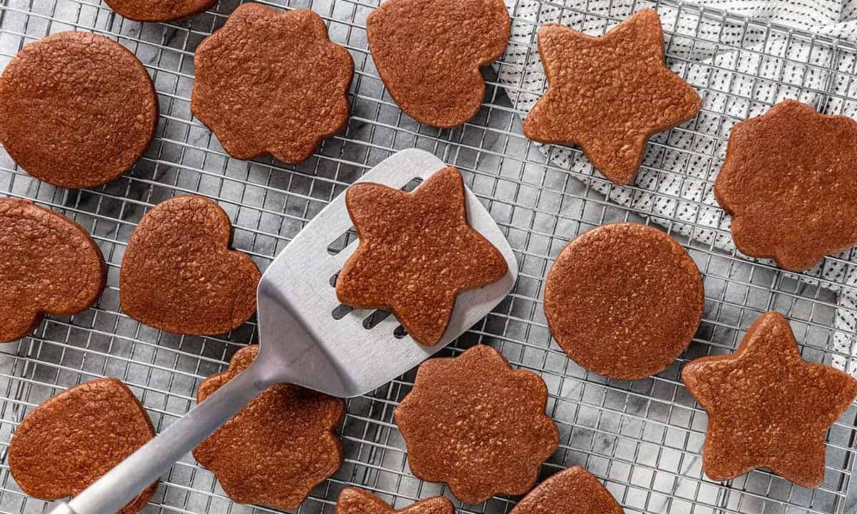 Bird's eye view of freshly baked chocolate sugar cookies laid on a wire cooling rack with a silver spatula picking up one star shaped cookie.