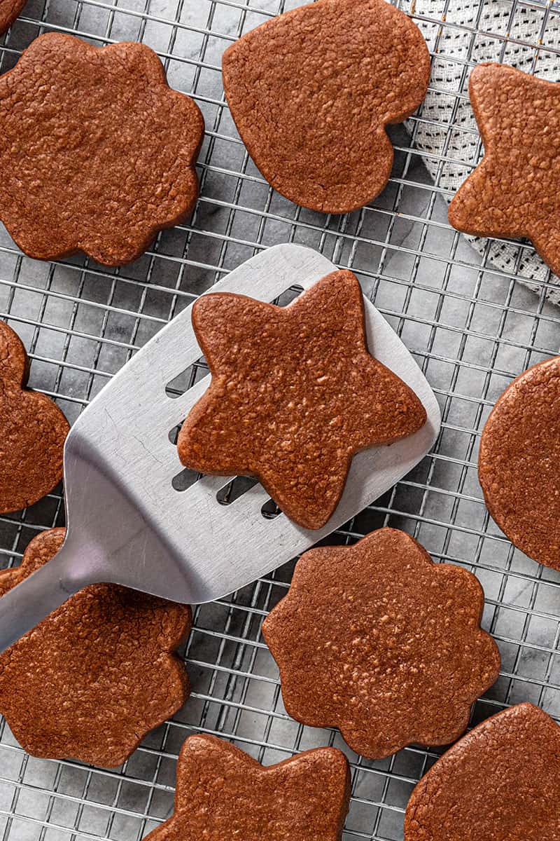 A spatula lifts a star shaped chocolate sugar cookie from many sugar cookies on a cooling rack.