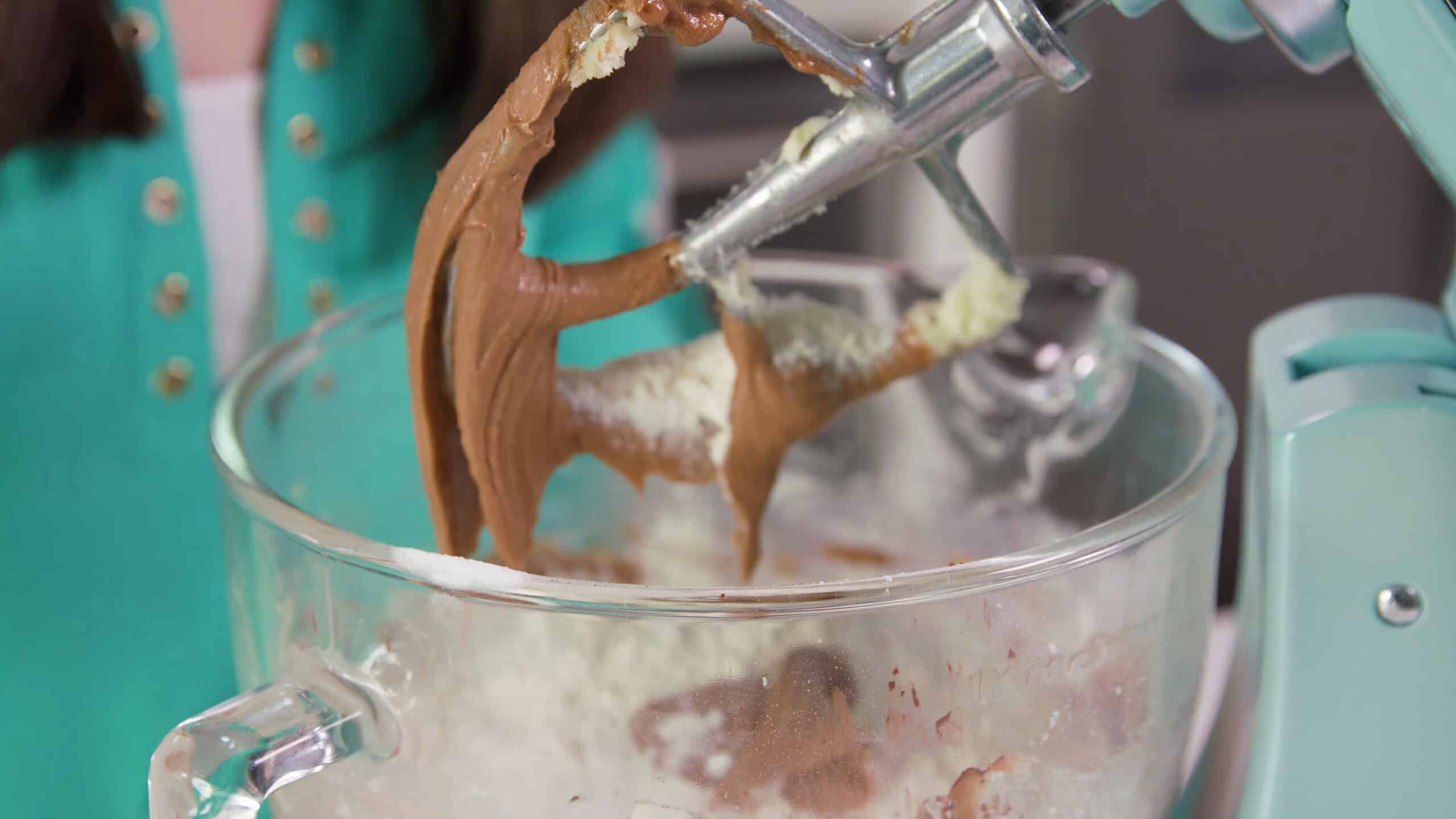 Angled view of metal paddle attachment from a countertop stand mixer with chocolate and butter mixture combined in a large clear glass mixing bowl.
