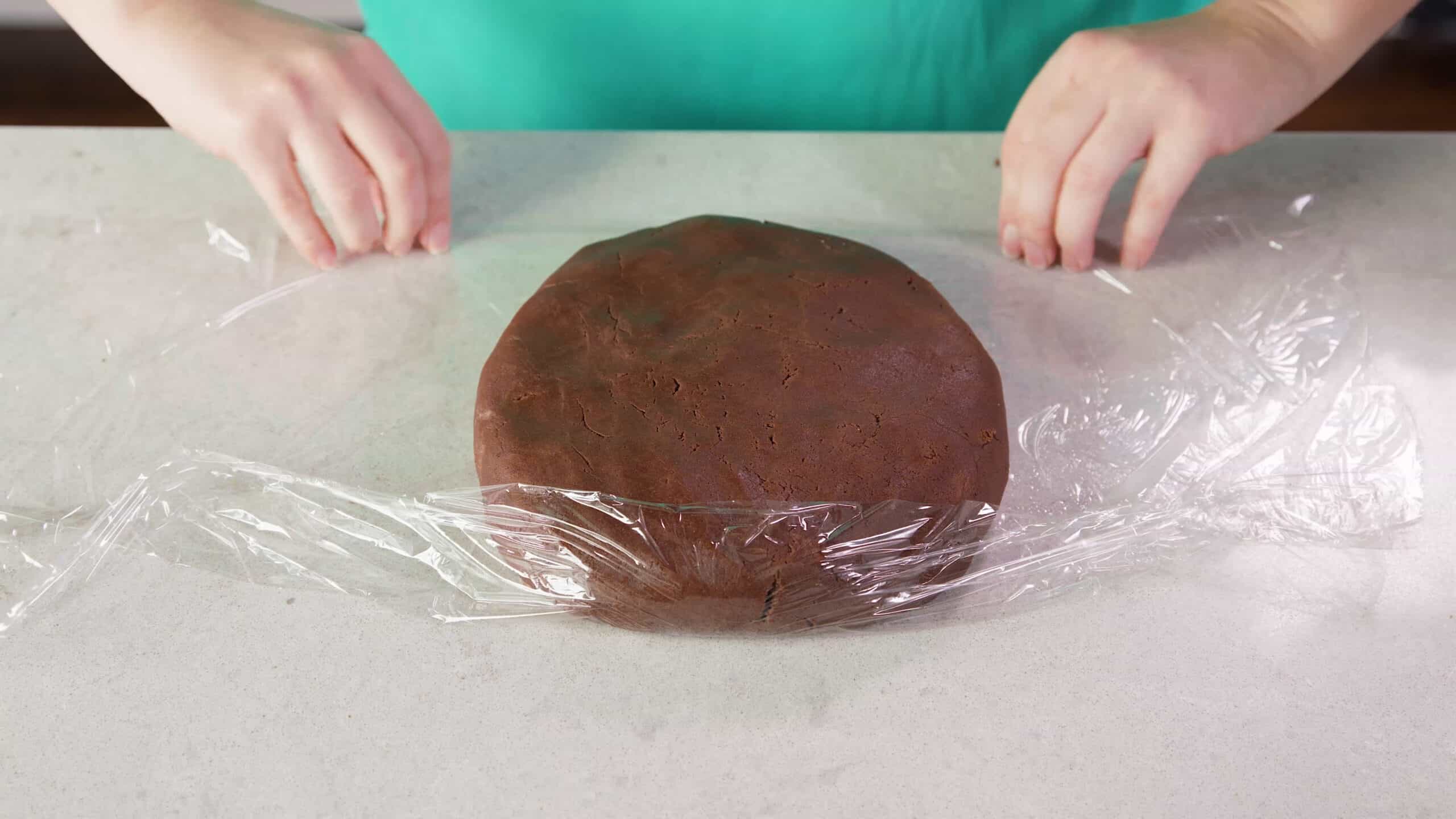 Overhead view of chocolate sugar cookie batter combined and shaped into a dough ball and wrapped in clear plastic wrap all on a clean marble countertop.