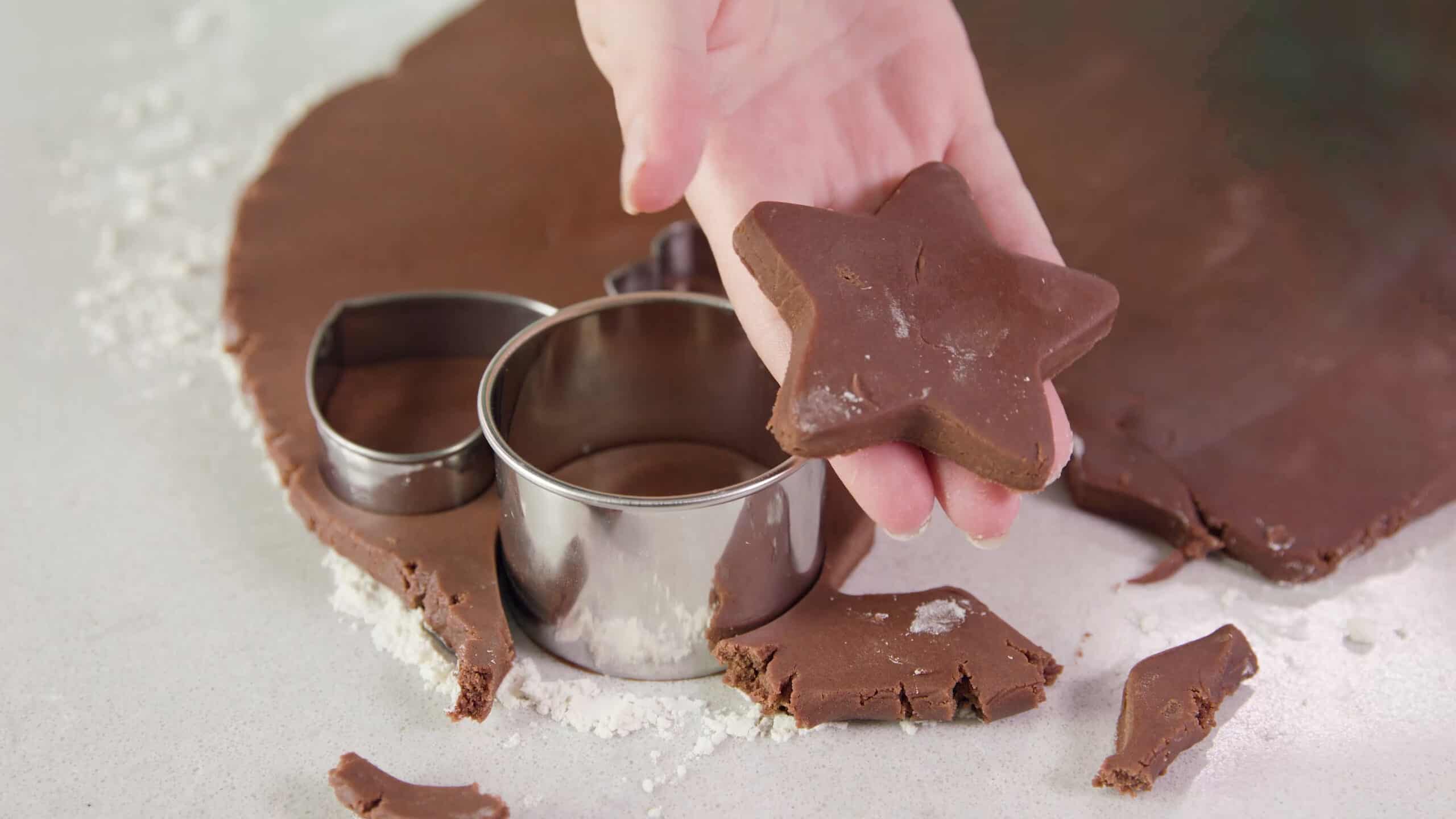 Angled view of a hand holding a star shaped chocolate sugar cookie after being punched from the flattened chilled dough using a metal cookie cutter, with the remaining flattened dough and other cookie cutters in the background.