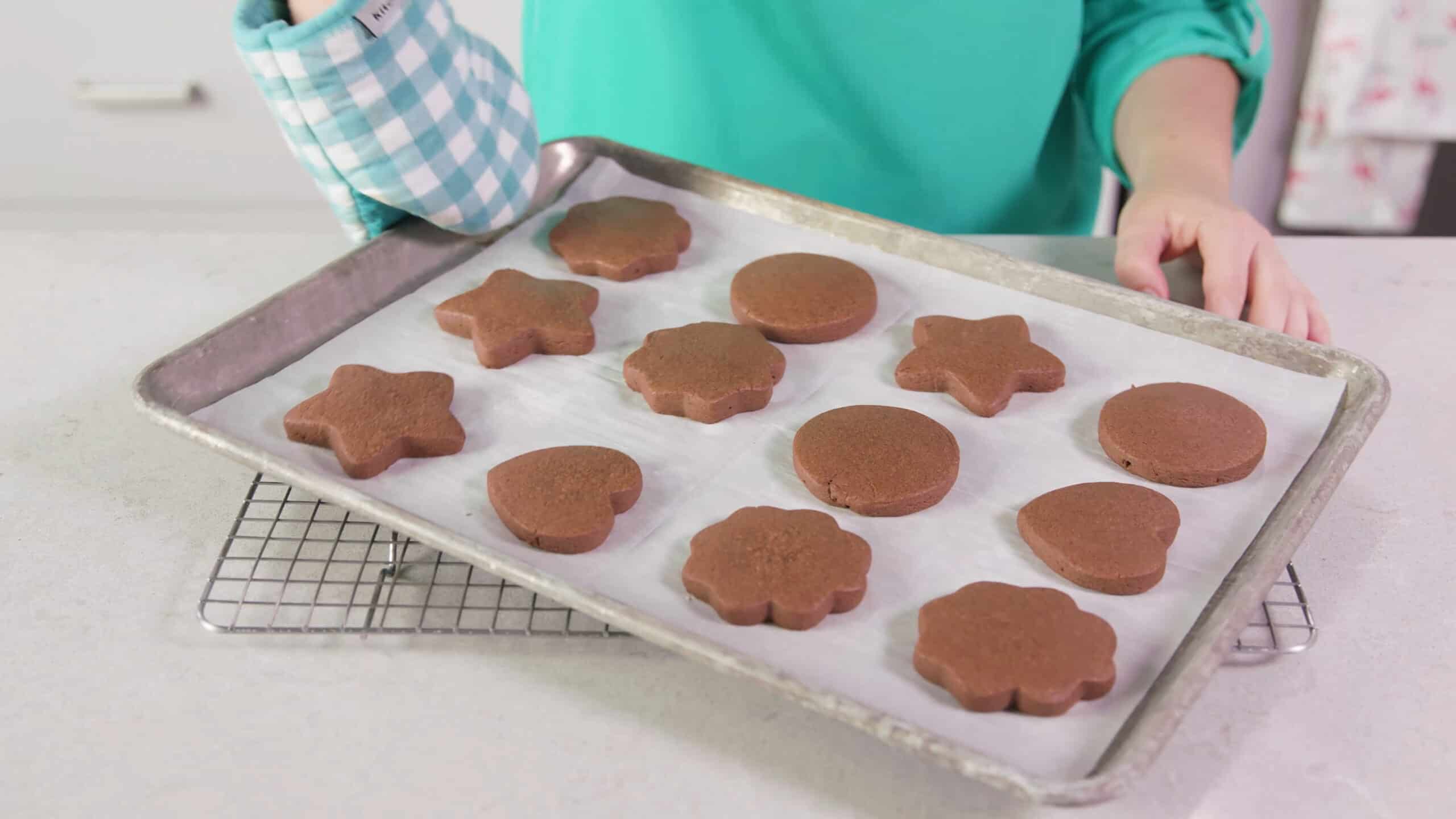 Angled view of metal baking sheet lined with parchment paper and covered with cooked chocolate sugar cookies of various shapes placed on a cooling rack that is on a marble countertop.