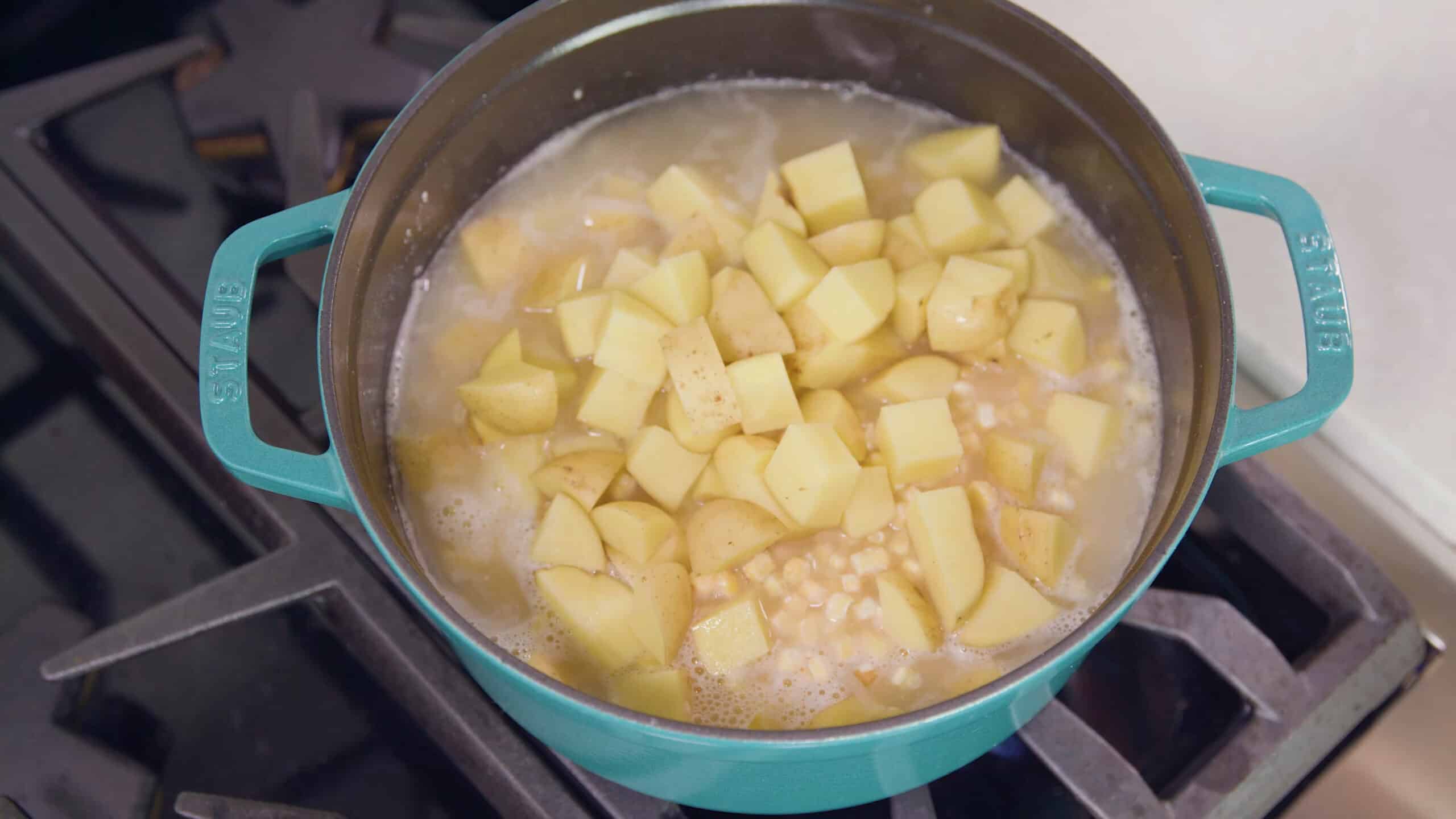 Overhead view of enamel coated cast iron pot filled with corn, onions, flour (for thickening), and Yukon gold potatoes, all on a stovetop burner.