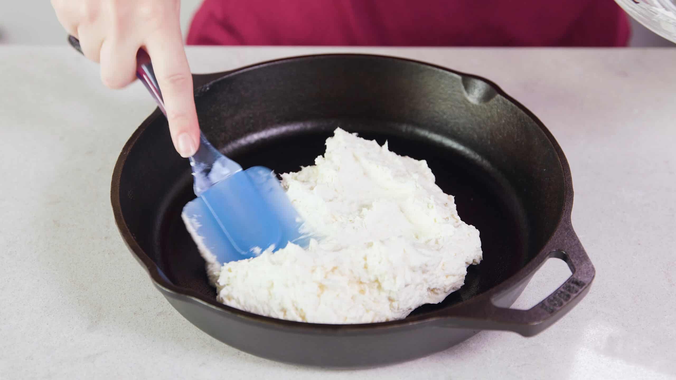 Angled view of cast iron skillet filled with cream cheese mixture and spread along the bottom of the pan by a plastic spatula all on a marble countertop.