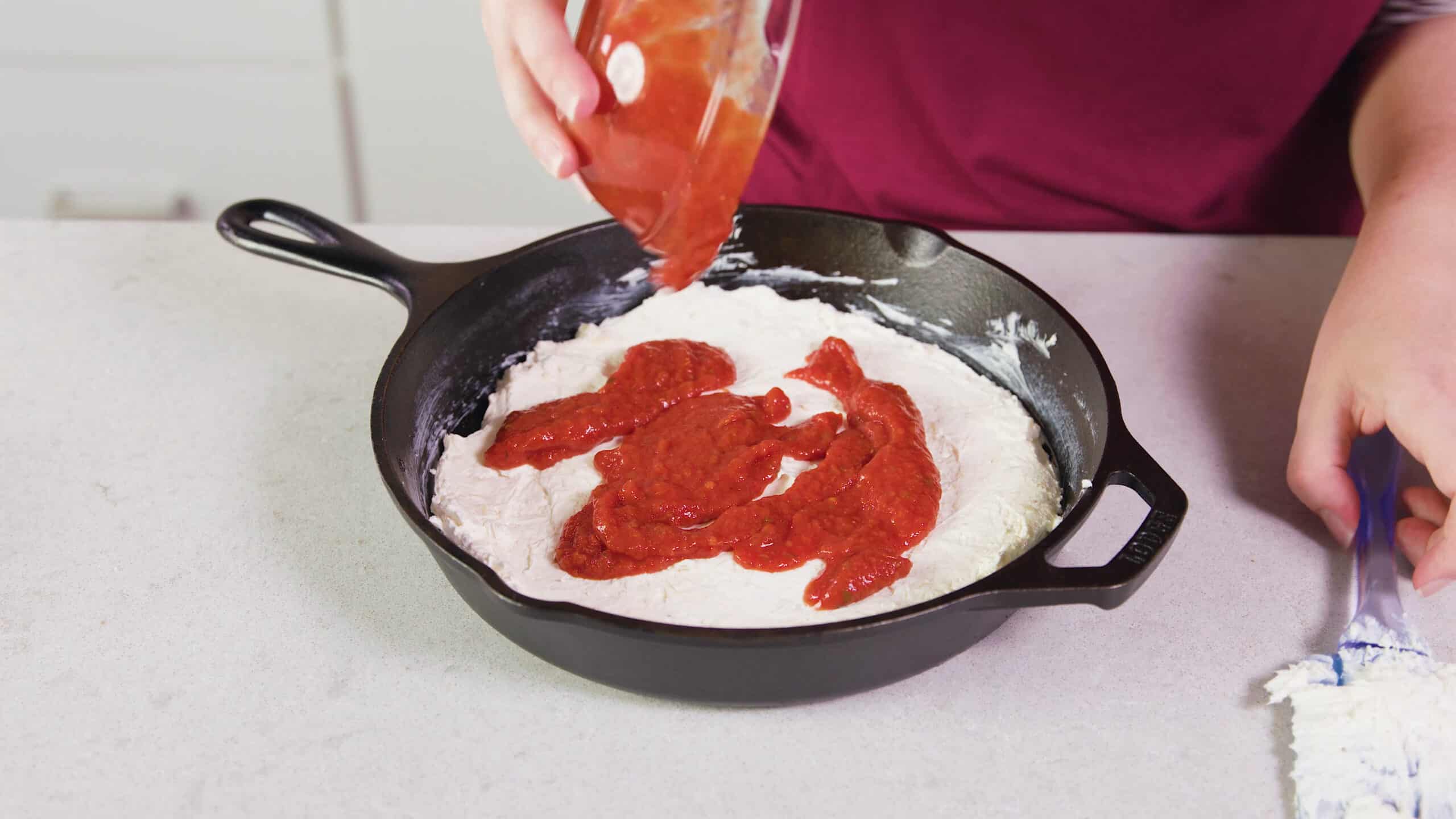 Angled view of cast iron skillet filled with cream cheese mixture lined along the bottom of the pan and marinara sauce poured on top from a clear glass mixing bowl.
