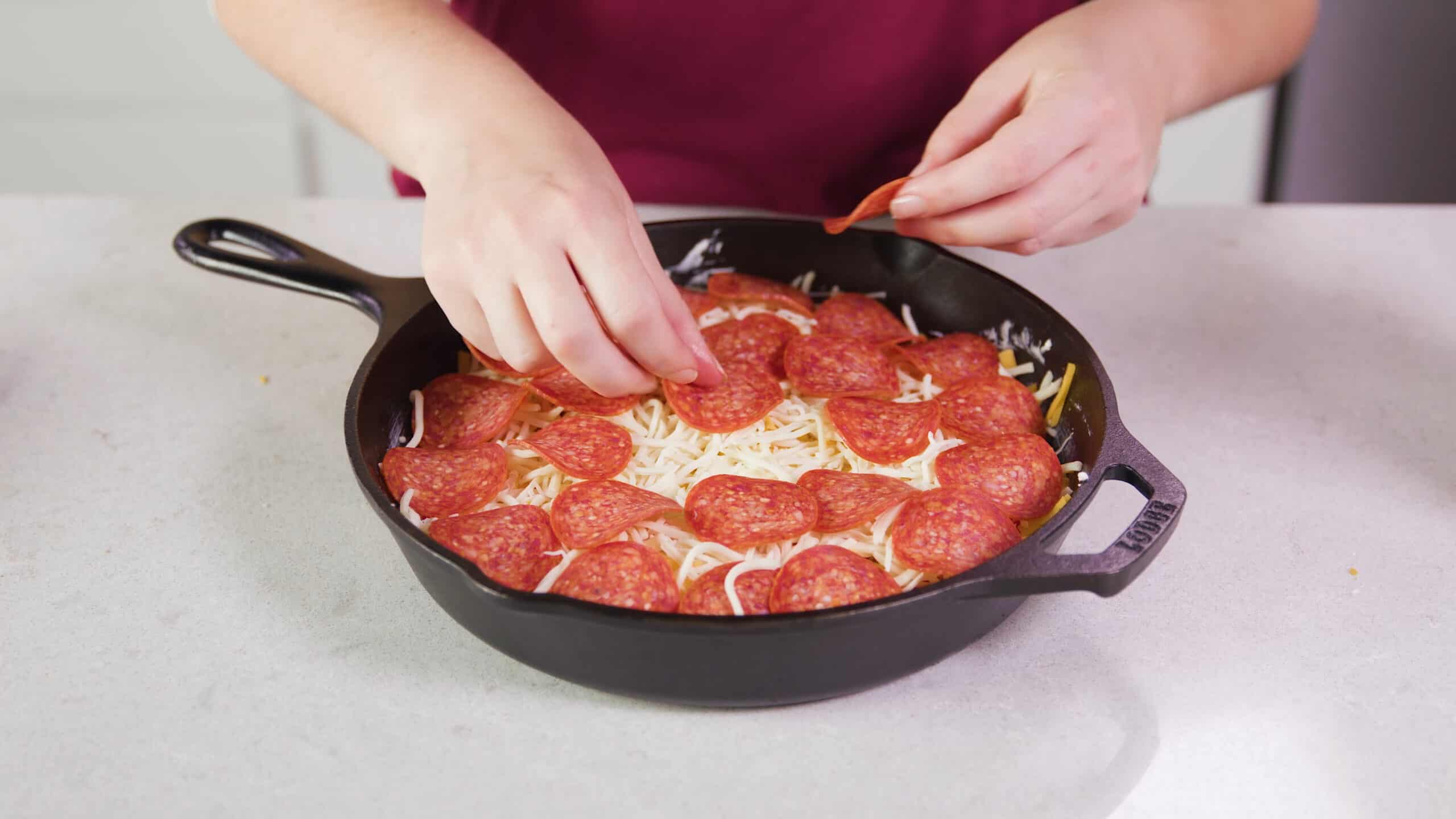 Angled view of cast iron skillet with cream cheese mixture lined with marinara sauce, shredded American and mozzarella cheeses and topped with slices of pepperoni.