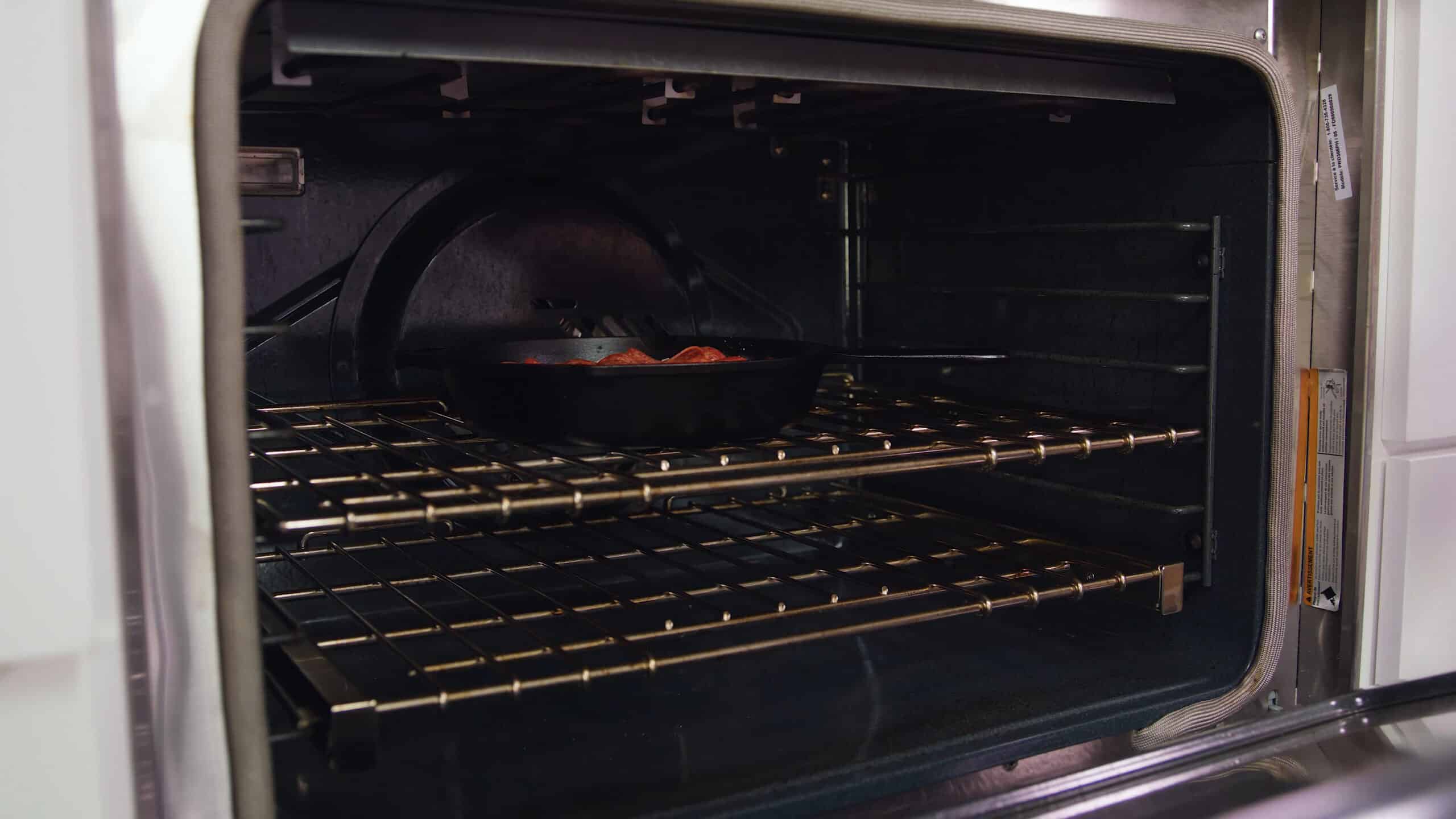 Angled view of an open oven with a cast iron skillet filled with ingredients for pizza dip placed on a metal rack in the middle of the oven.