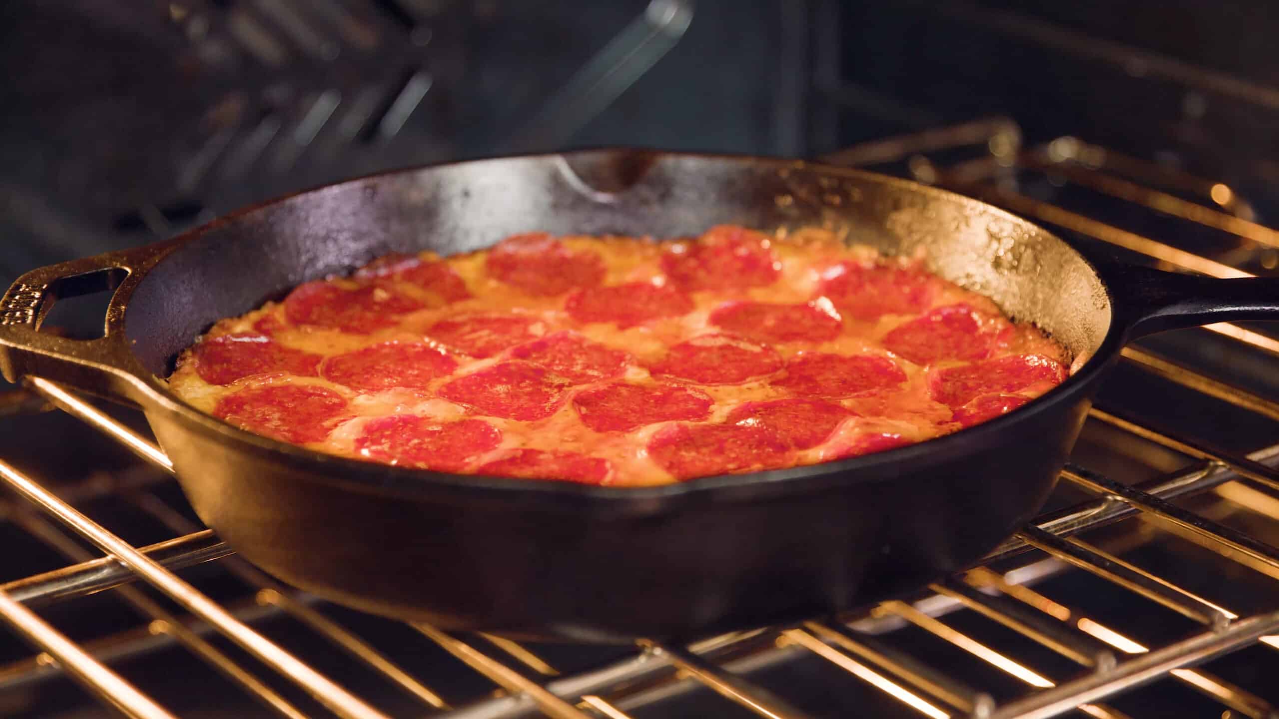 Close-up view of an open oven with a cast iron skillet filled with bubbling and sizzling pizza dip inside all on a metal rack in the middle of the oven.