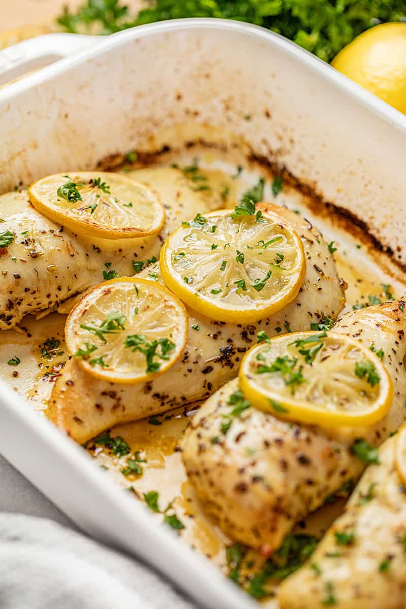 Close up view of Baked Chicken Breasts in a white pan topped with lemon slices and parsley.