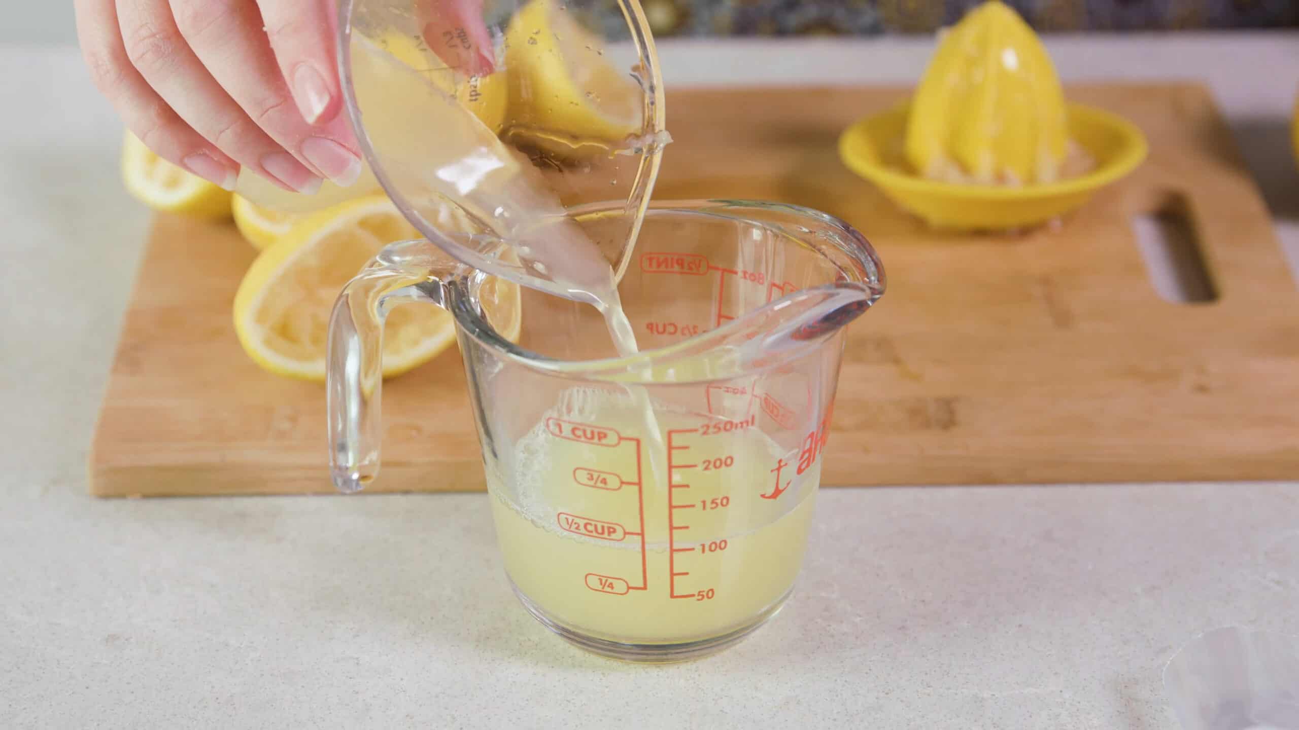 Angled view of clear glass measuring cup being filled with lemon juice from a hand held juicing cup, with a wood cutting board and lemon slices in the background.